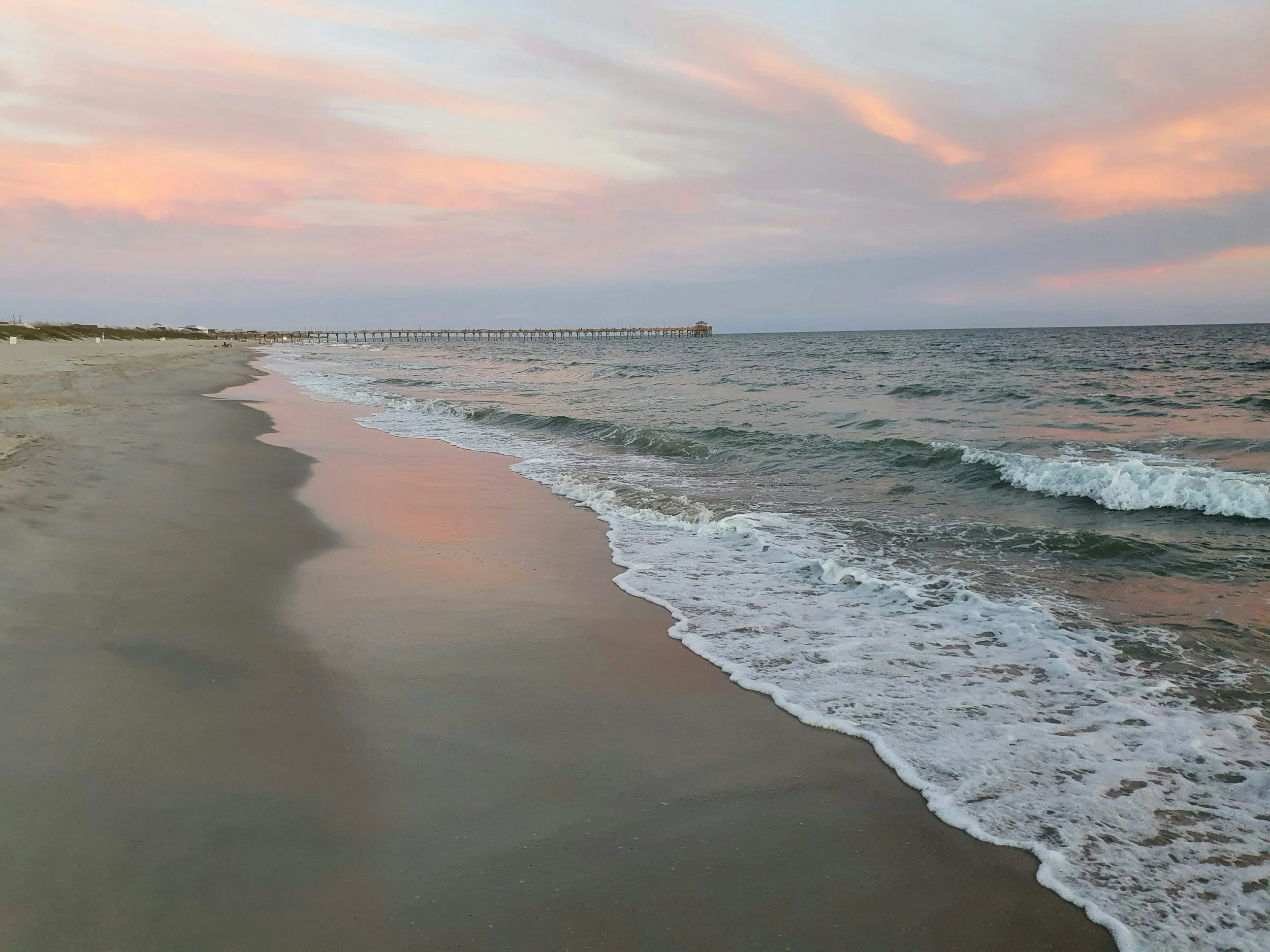 A sandy beach with gentle waves reaching the shore, a pier extending into the ocean, under a sky with pink and orange hues of a setting or rising sun.