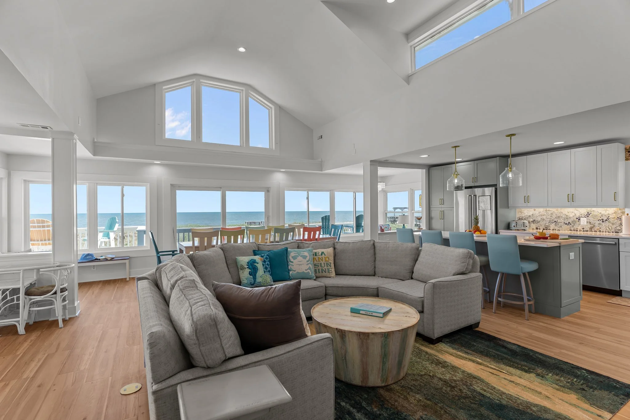 Living room with large windows, ocean view, white walls, gray sofa with colorful pillows, wooden coffee table, and open kitchen with light gray cabinets, bar stools, and pendant lights.