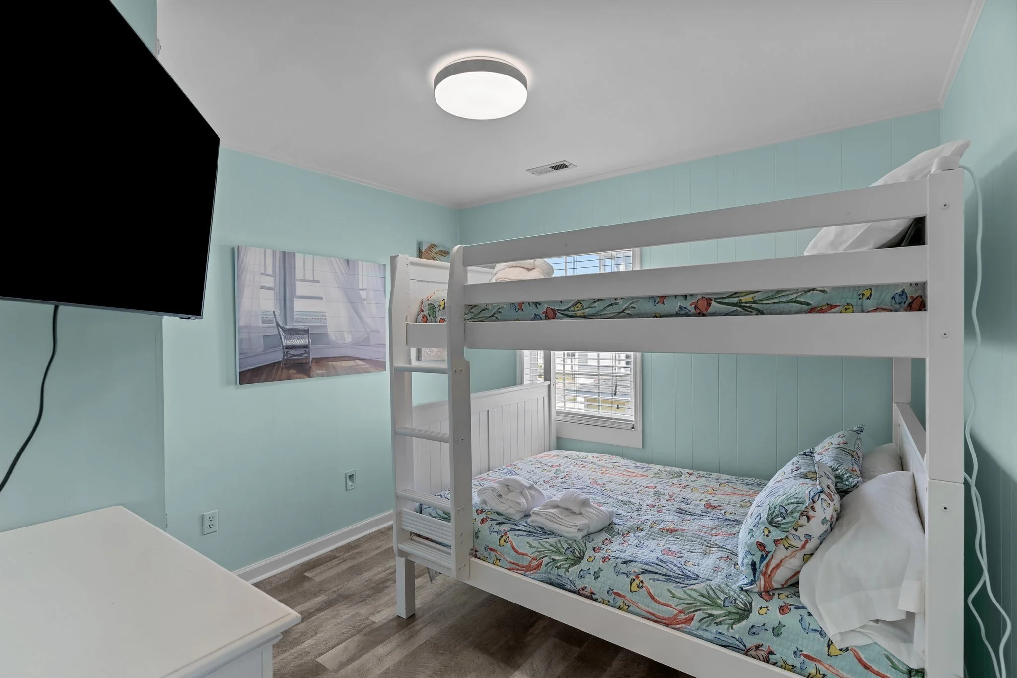 Bedroom with light blue walls, a white bunk bed with colorful patterned sheets, a wall-mounted television, and a window with white blinds.