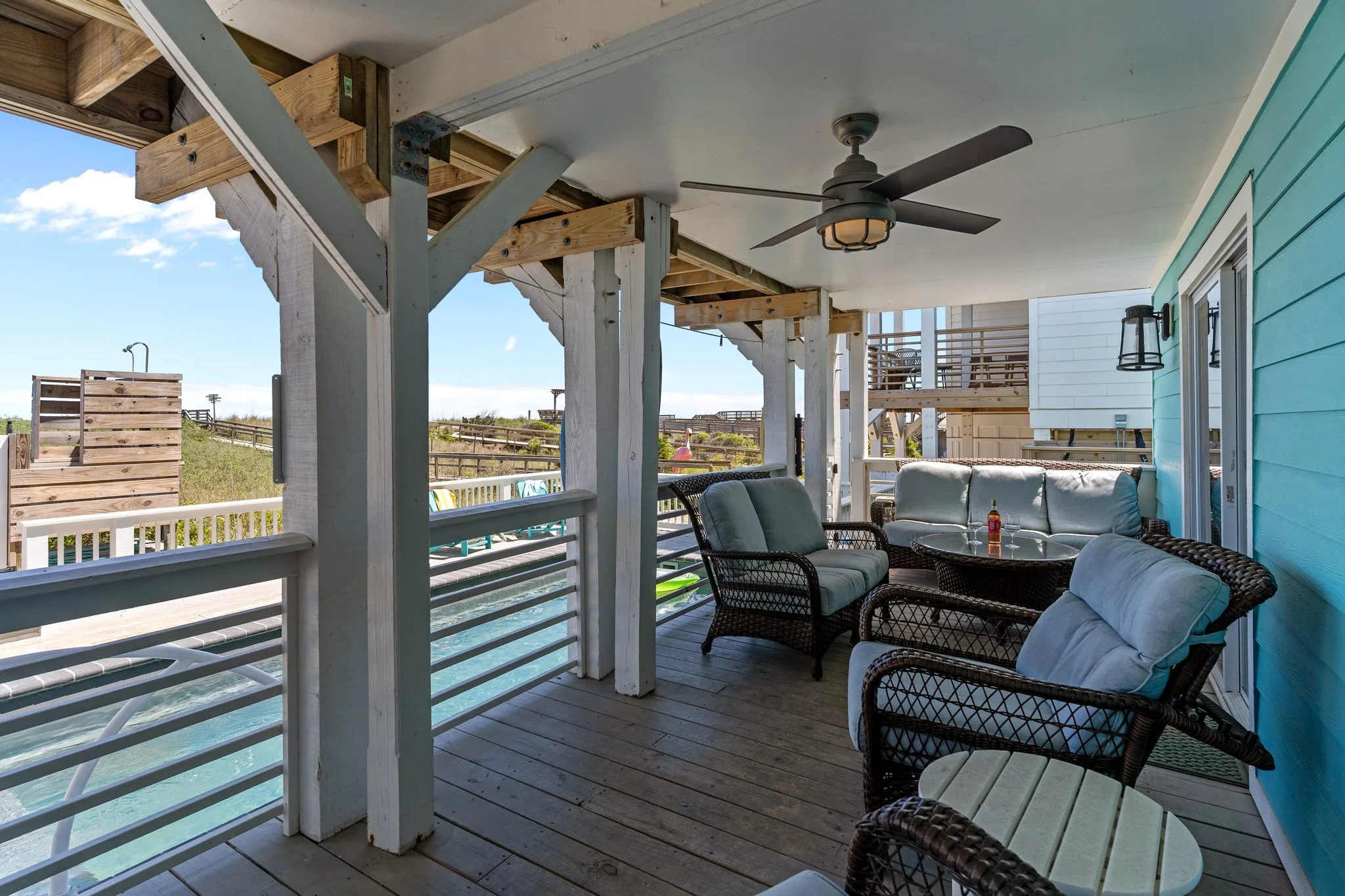 A covered patio with wicker furniture, including a loveseat and chairs, decorated with cushions. There is a ceiling fan with lights, and the porch overlooks a waterfront with a wooden railing, a paddleboard, and a bright blue house wall.