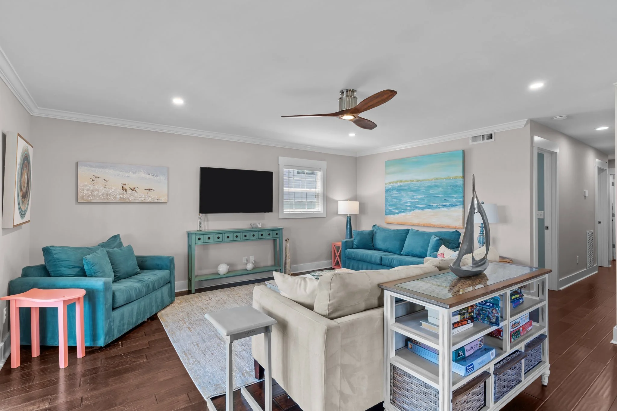 Living room with blue sofas, artwork, a flat-screen TV, a lamp, a ceiling fan, and a console table with decorative items.