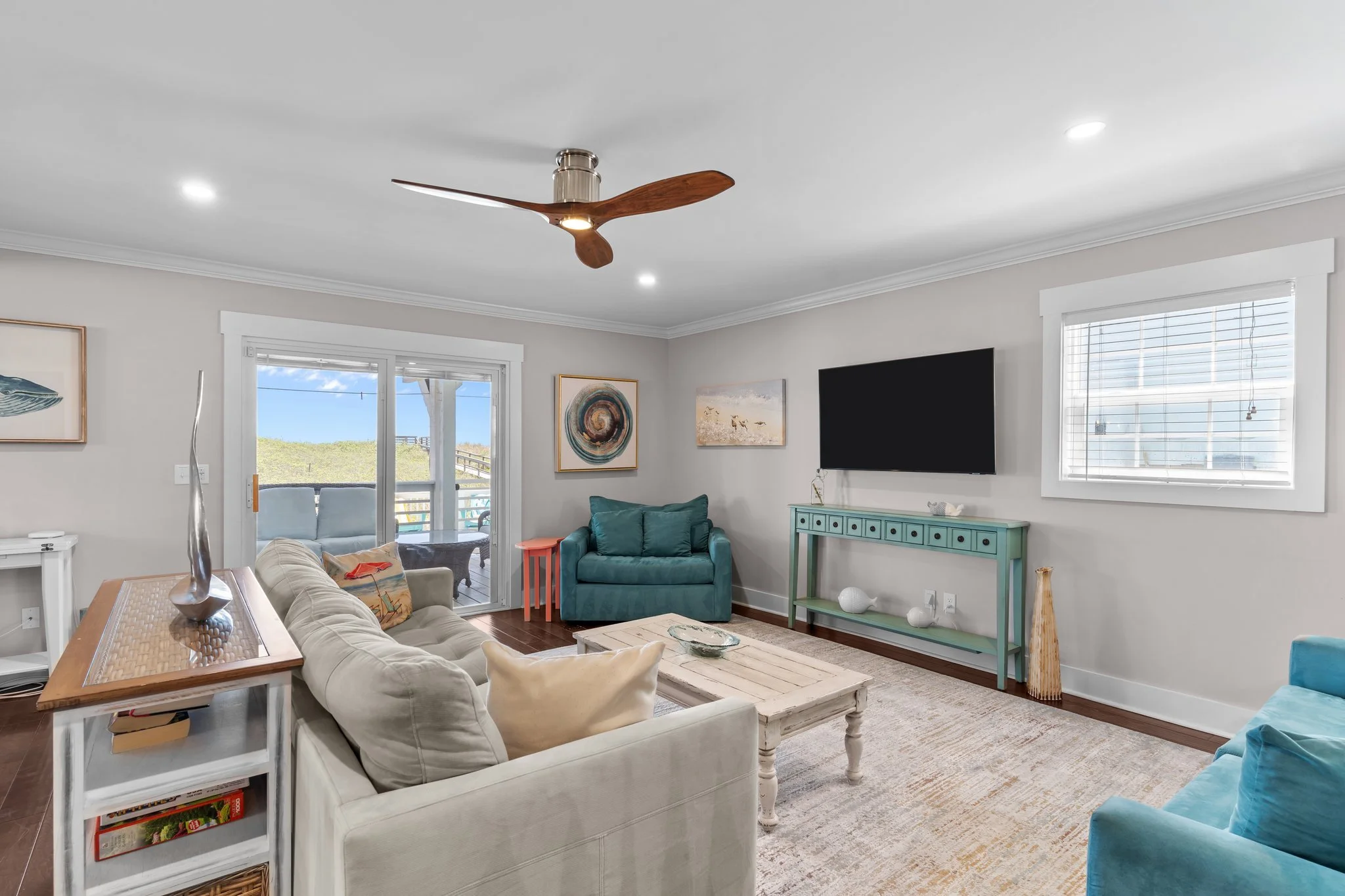 Living room with white sofa, green armchair, wooden coffee table, wall art, flat-screen TV, ceiling fan, large windows with blinds, and decorations.
