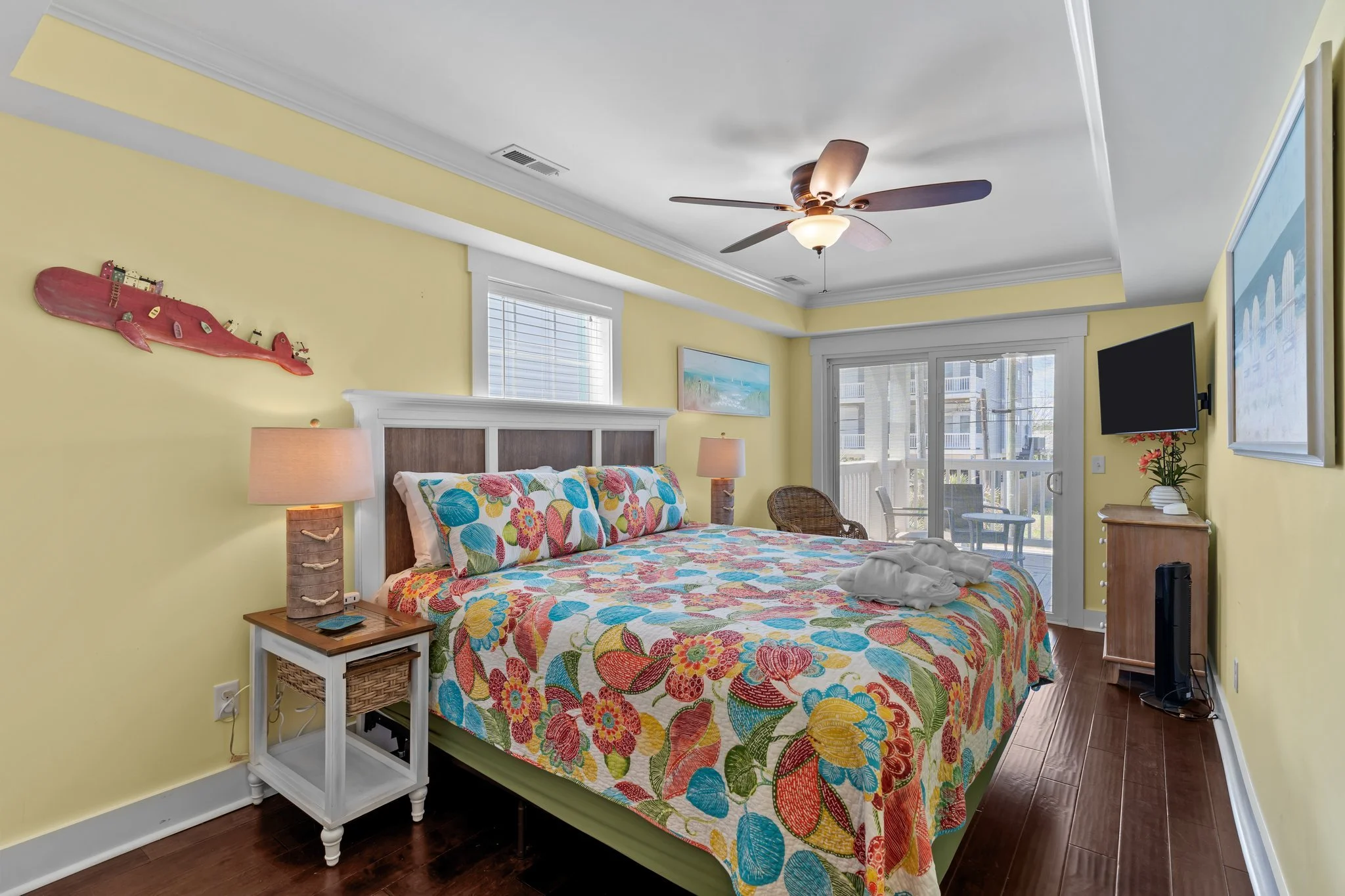 A cozy bedroom with yellow walls, a colorful floral bedspread, and a wooden headboard. There is a white nightstand with a woven lamp, a window with blinds, and a sliding glass door leading to a balcony. A ceiling fan and wall art are also visible.