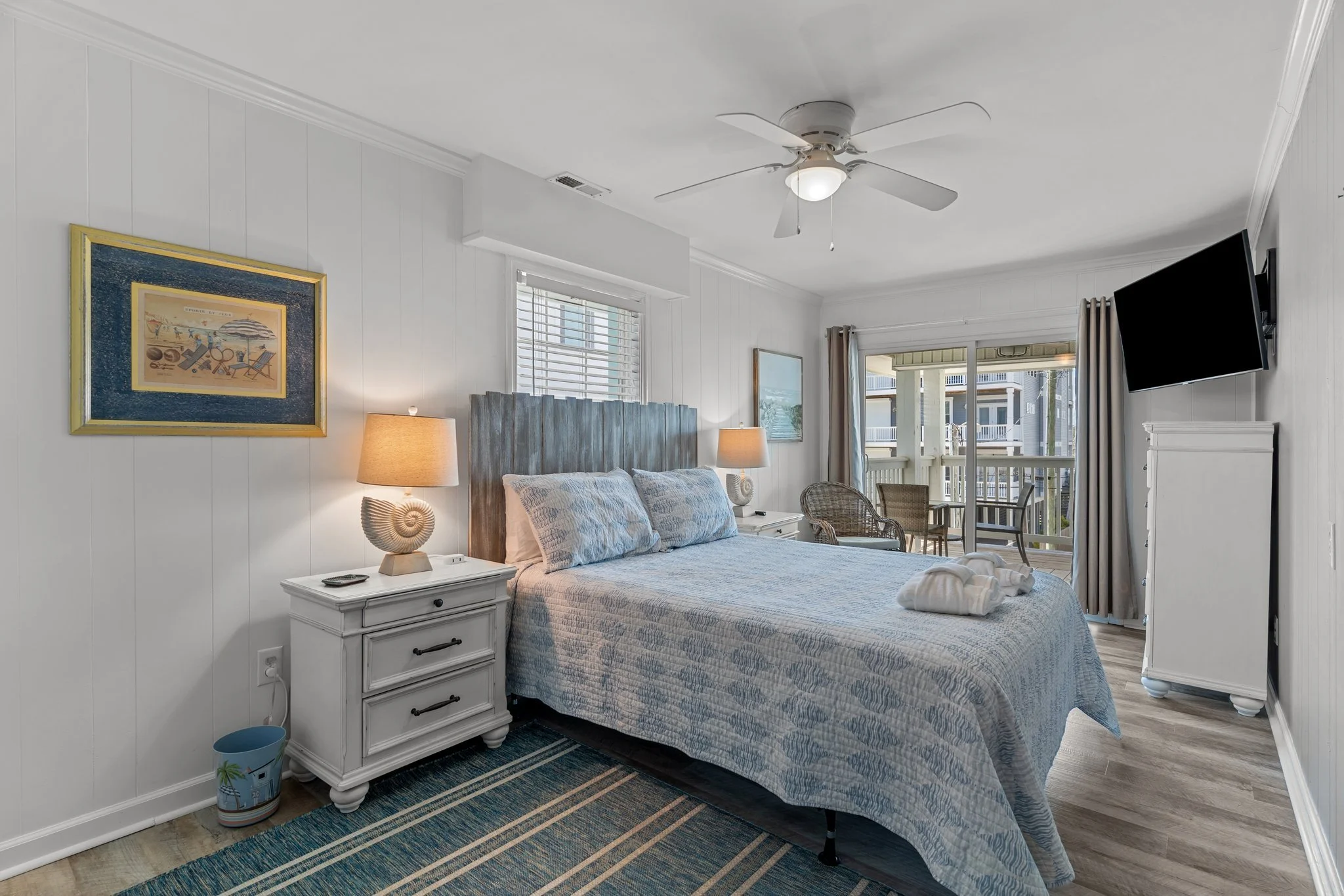 Bright hotel bedroom with white walls, a large bed with a quilted comforter, a wooden headboard, two nightstands with lamps, a framed picture on the wall, and a sliding glass door with curtains leading to a balcony.