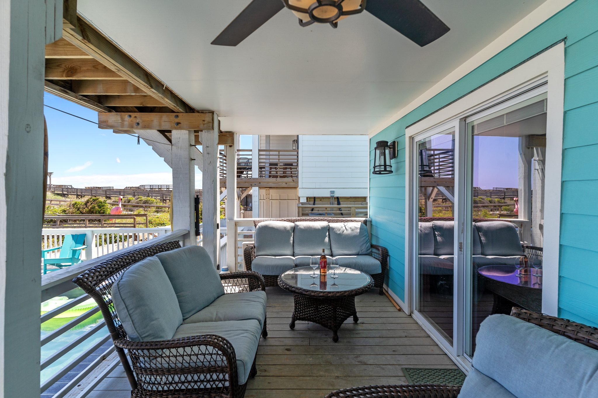 A porch with outdoor wicker furniture including chairs and a loveseat with light cushions, a round glass-top coffee table with drink bottles, sliding glass door, turquoise house wall, ceiling fan, outdoor light fixture, and a view of a fenced yard wi