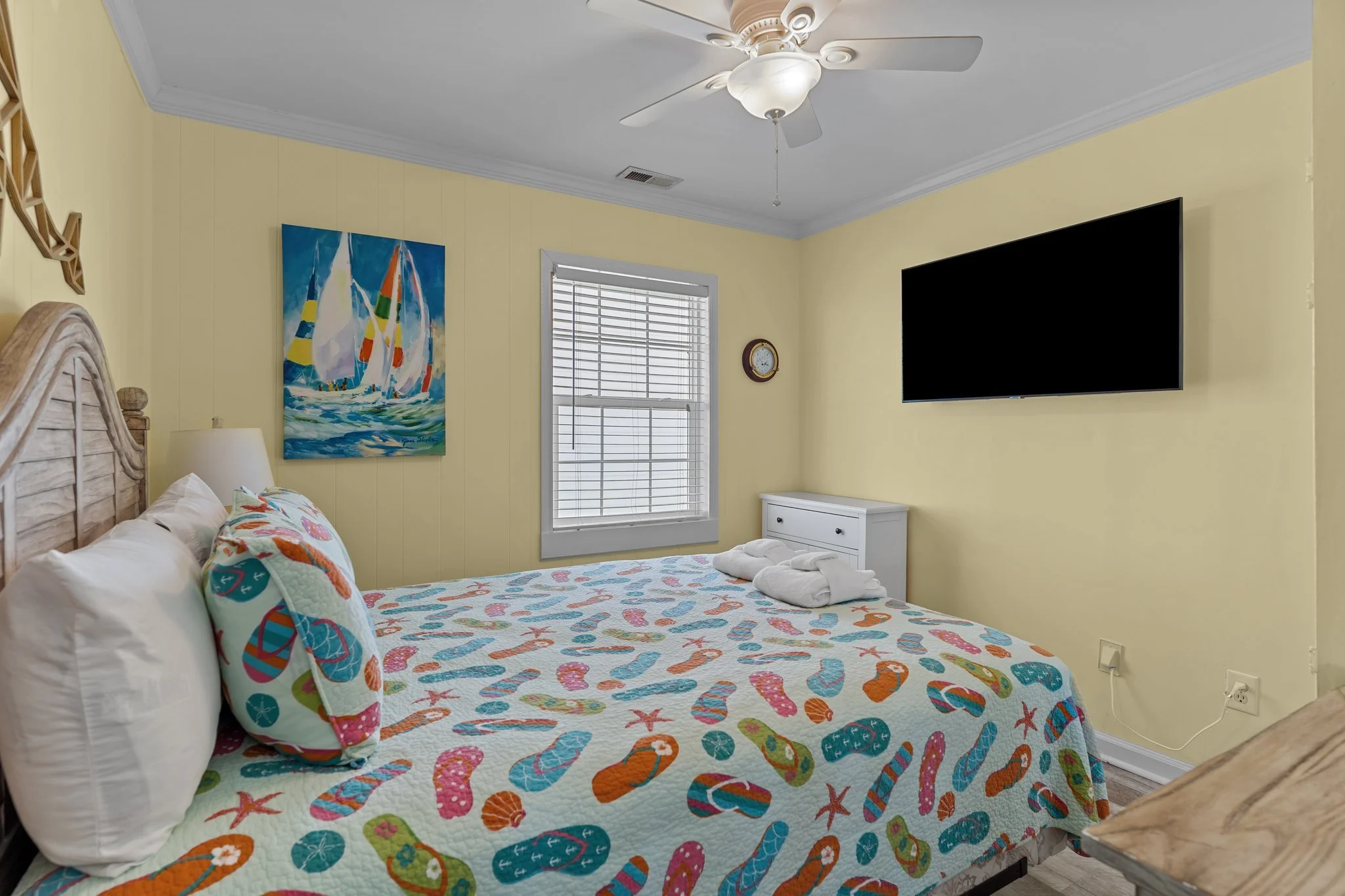 A cozy bedroom with a yellow painted wall, a bed with a colorful tropical-themed quilt, white pillows, and a seashell pillow. There is a window with blinds, a painting of sailboats, a small clock, a white dresser, a ceiling fan with lights, and a mou