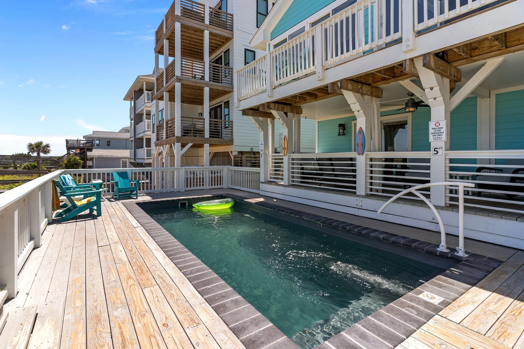 Deck with small swimming pool, blue and green chairs, and a colorful inflatable float, with multi-story beach house in the background under a clear blue sky.
