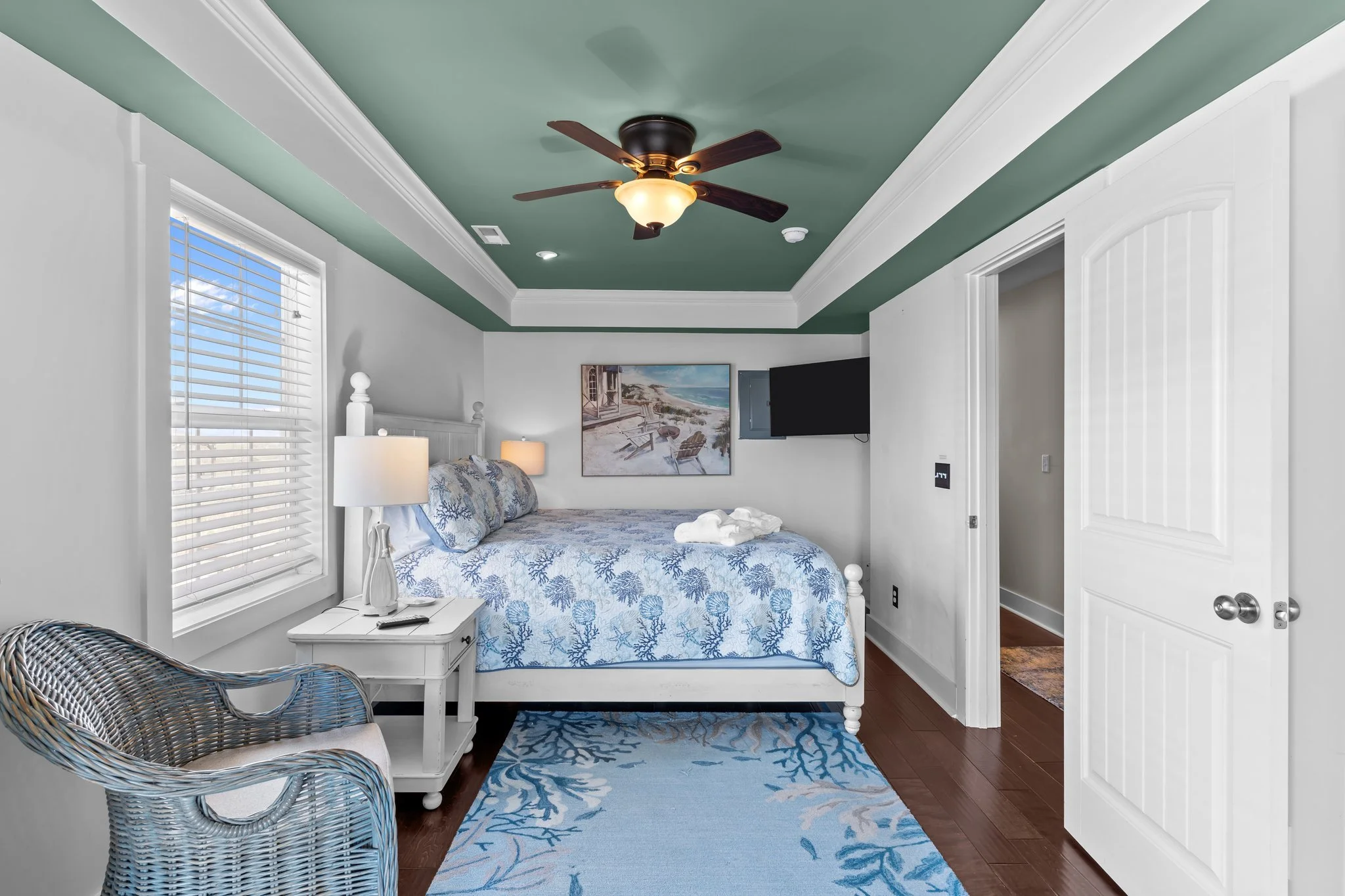 A bedroom with a white bed, blue and white ocean-themed bedding, and a wicker chair. The ceiling is painted green with white crown molding and a ceiling fan. There is a window with white blinds and a painting of a beach scene on the wall.