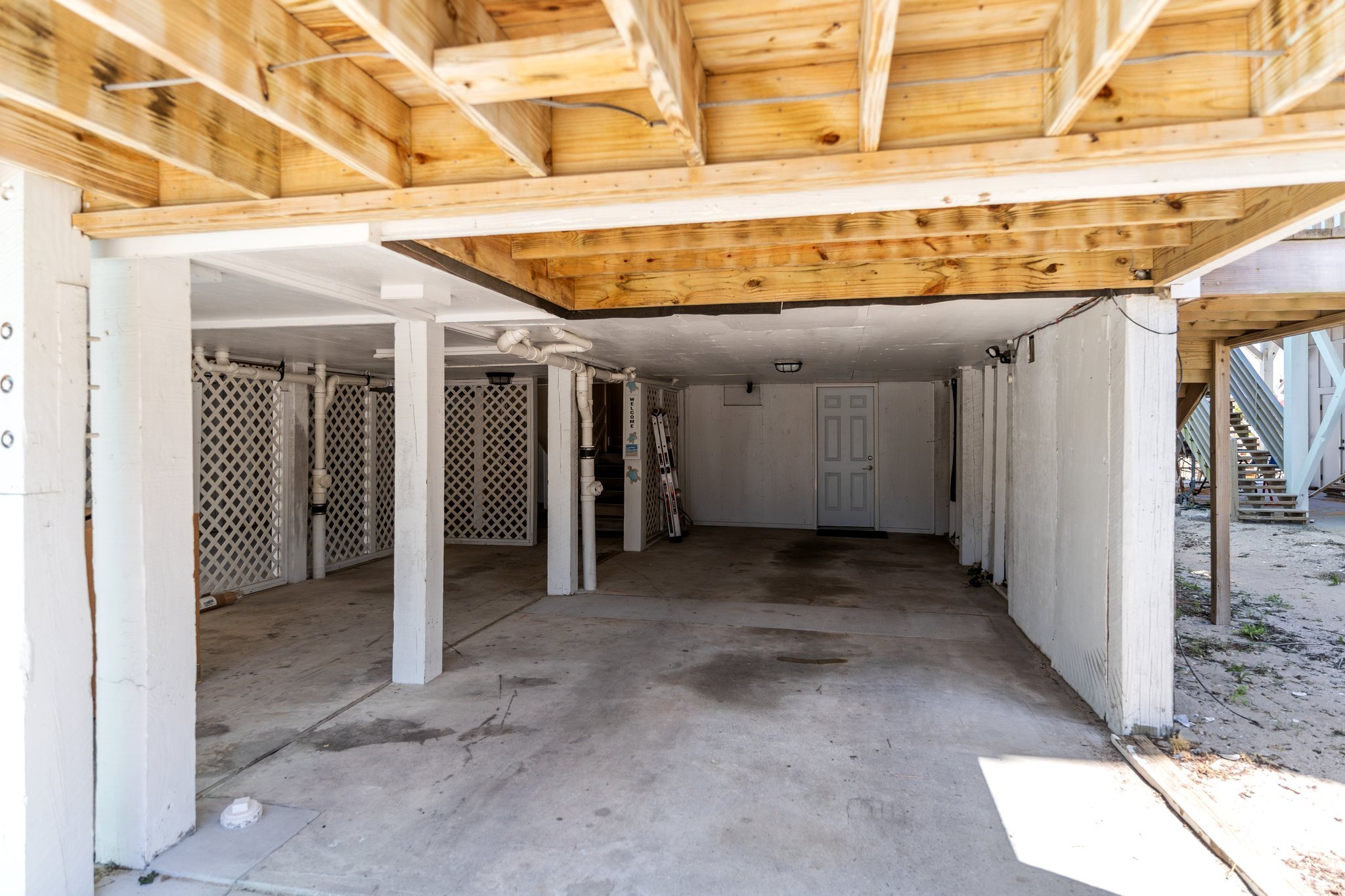 Under construction or renovation space with exposed wooden framing on the ceiling, concrete floor, and white painted support beams. The area appears to be a garage or basement, with some white lattice panels and PVC pipes visible along the walls.