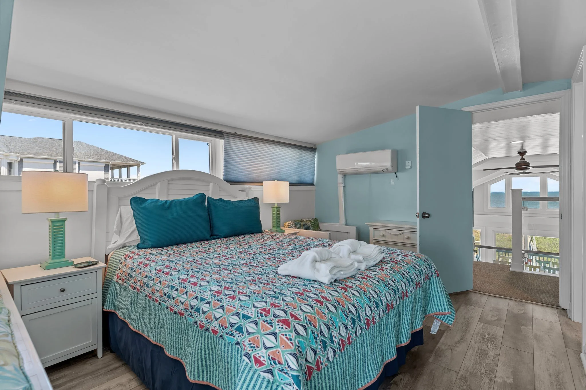 A cozy bedroom with a large bed covered in a colorful patterned quilt, two blue pillows, and a folded white towel on top. There are two bedside lamps with green bases on white nightstands. The room has large windows with shades, allowing natural ligh