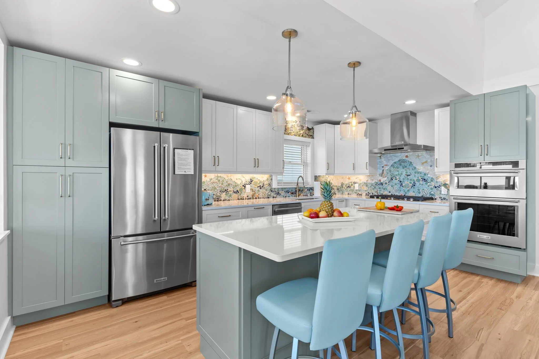 A modern kitchen with blue cabinets, stainless steel appliances including a refrigerator and oven, a white marble island with blue chairs, and colorful fruit and vegetables on the counter and island. There are two hanging pendant lights and a mosaic 