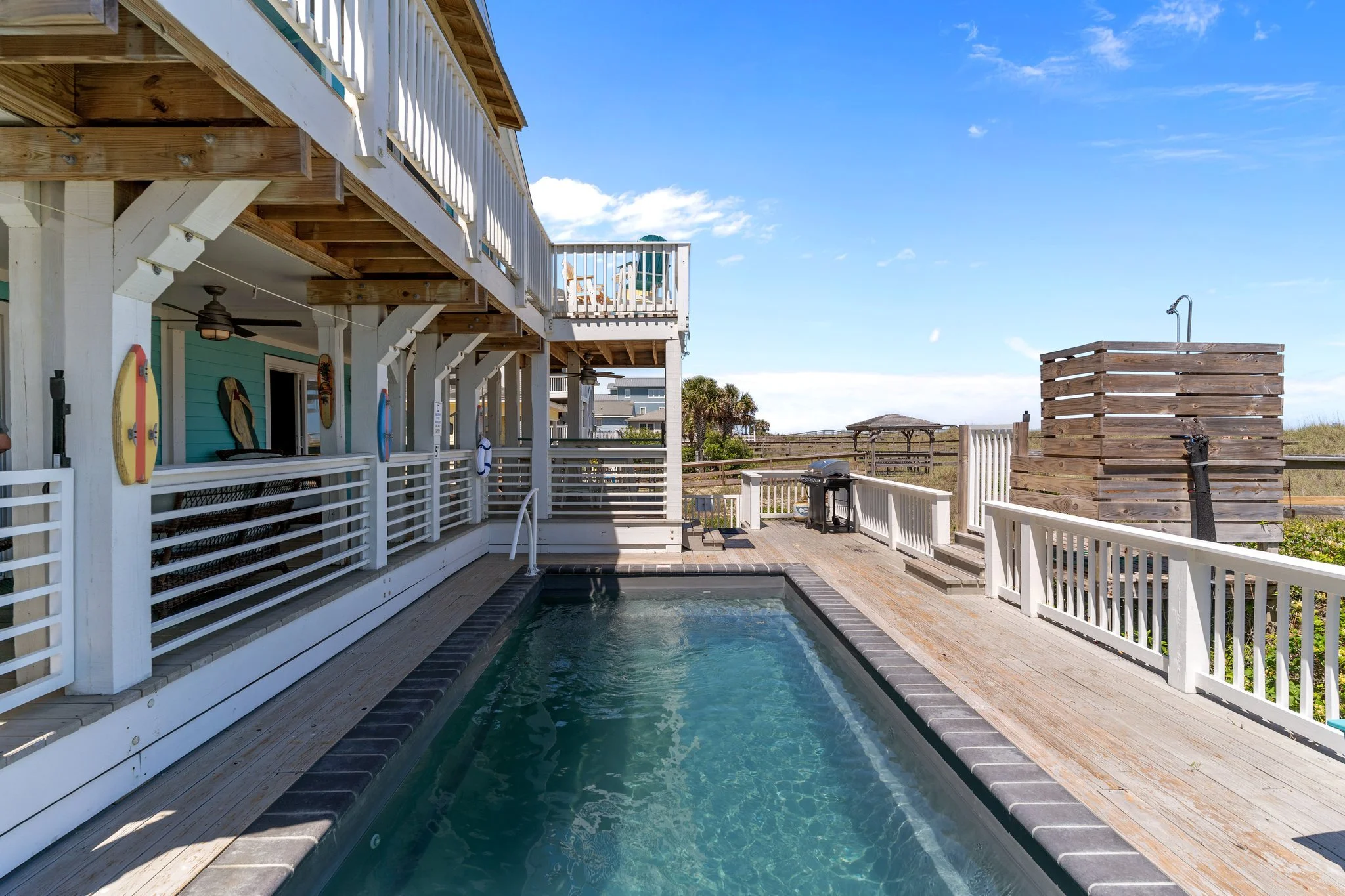 A backyard with a small swimming pool, white fencing, and a wooden deck. There is a two-story house with a balcony, outdoor furniture, and surfboards mounted on the wall. The sky is clear with some clouds, and there is a wooden shower and some palm t