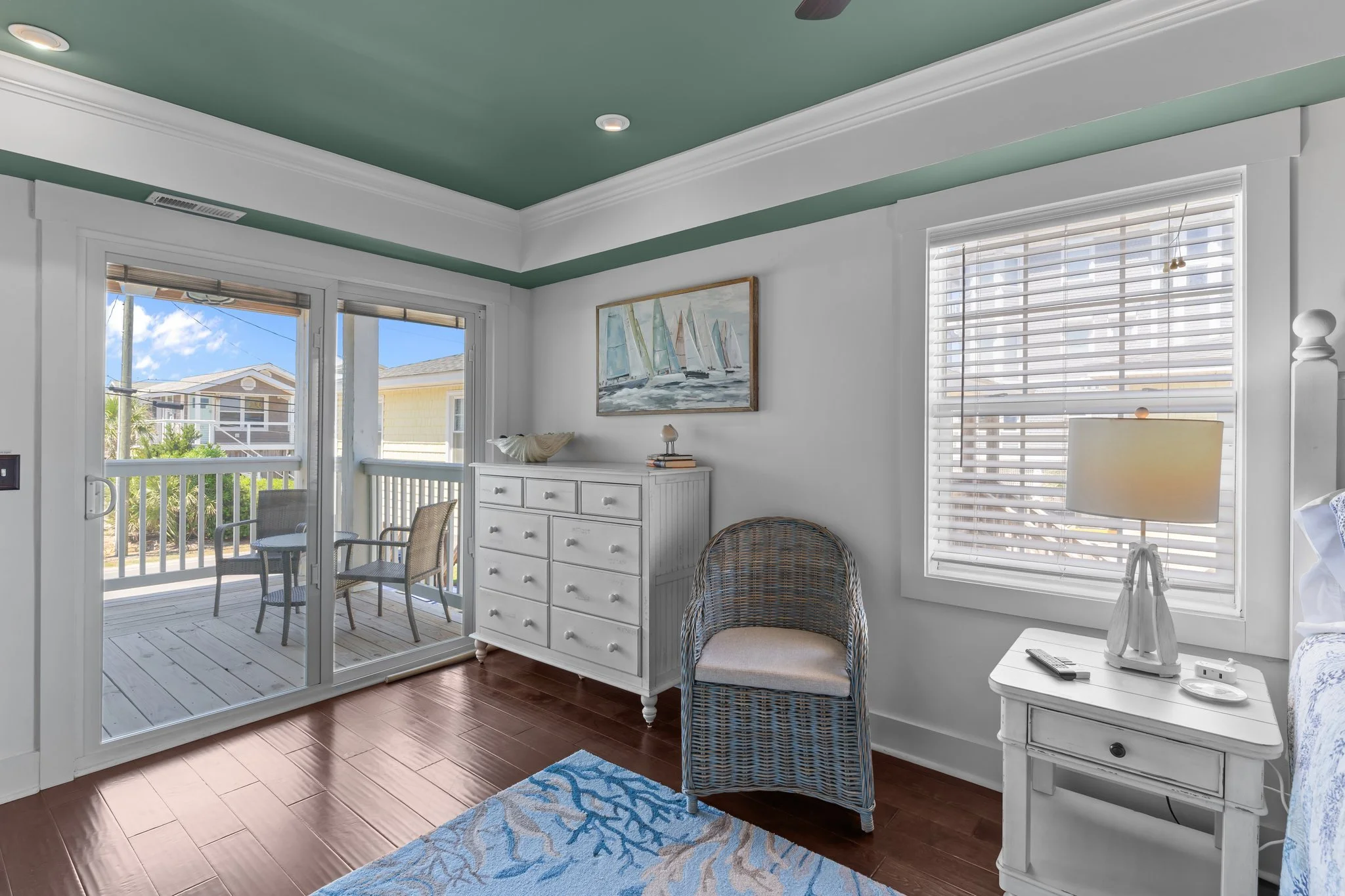 A cozy room with white walls and wooden floors, featuring a sliding glass door leading to a balcony, a wicker chair, a white dresser with artwork of sailboats hanging above, and a window with shades, allowing natural light to illuminate the space.