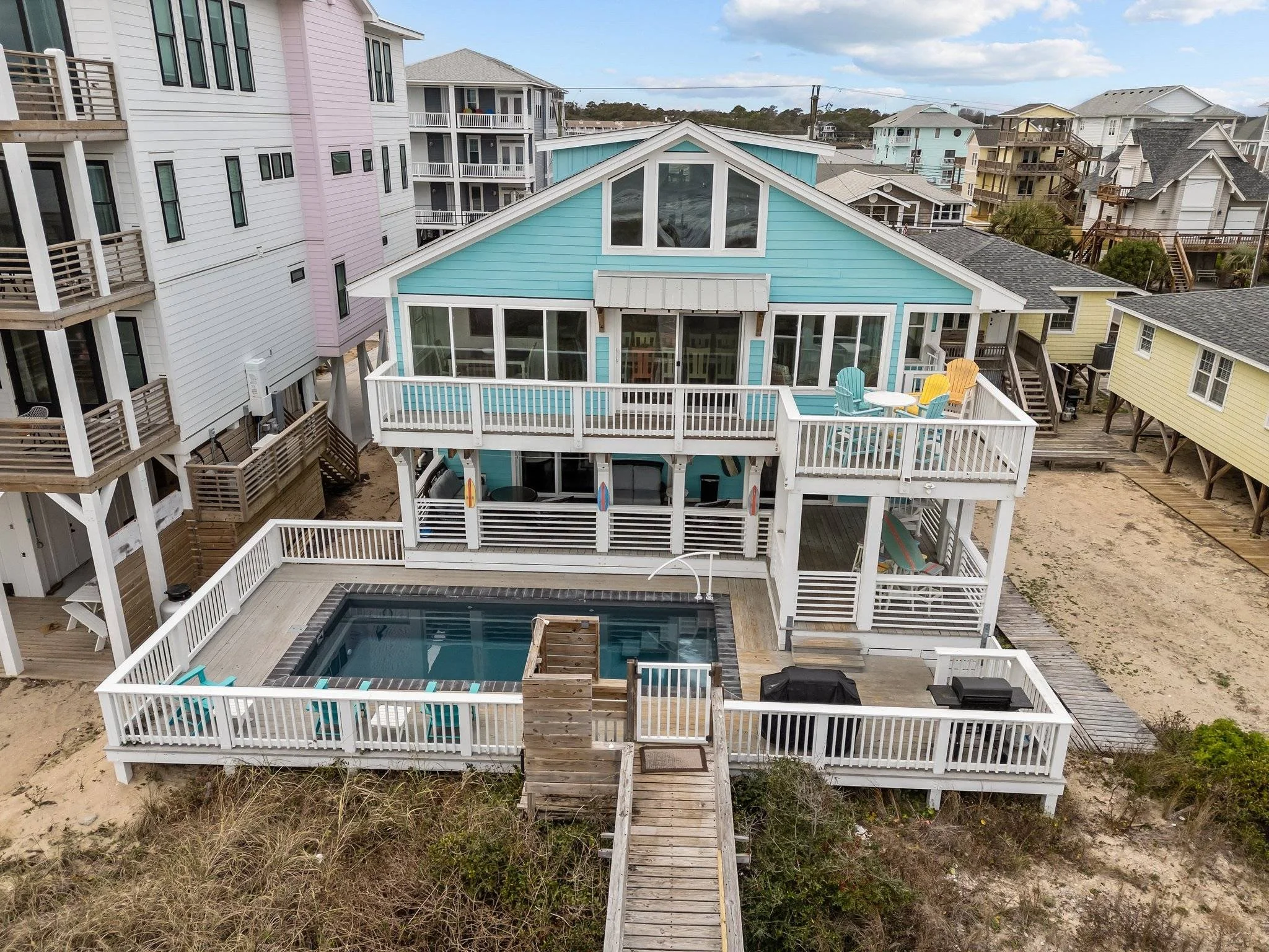 Aerial view of a coastal neighborhood featuring colorful beach houses with decks, stairs, and a swimming pool surrounded by sandy ground and grassy dunes.
