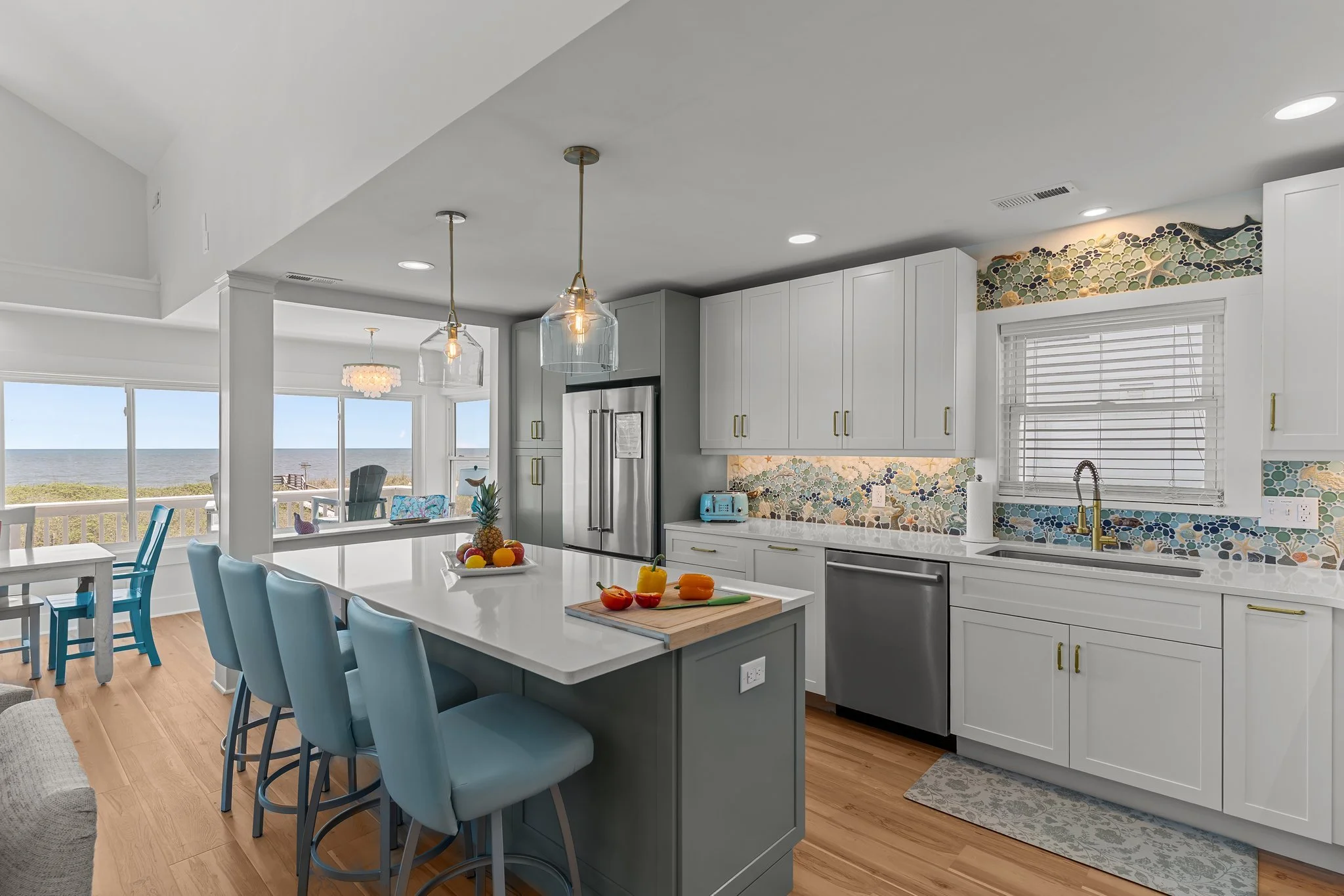 Bright kitchen with white cabinets, a large island with blue chairs, and a view of the ocean through large windows.