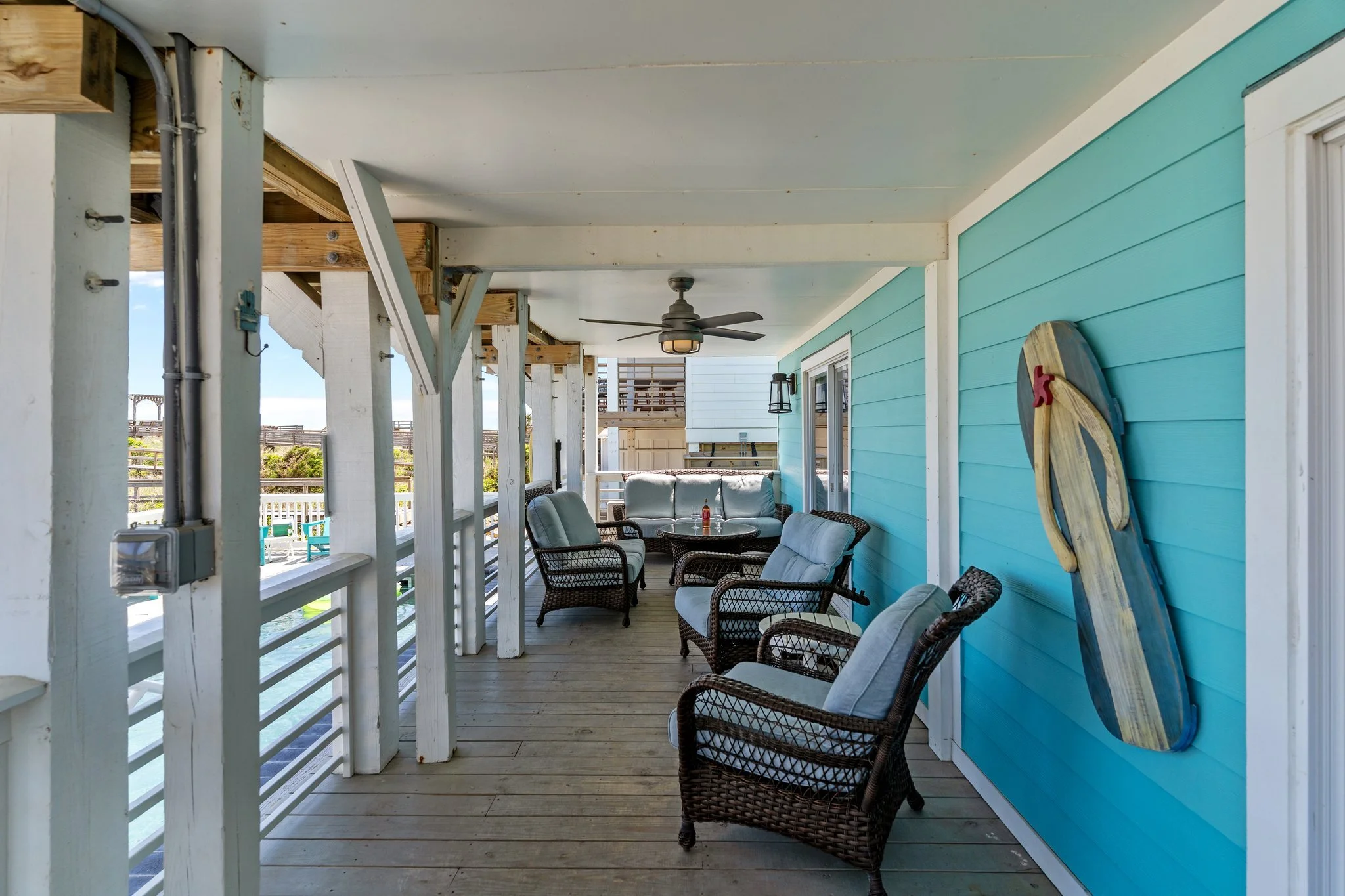 Porch area with turquoise siding, wicker chairs with cushions, ceiling fan, and a decorative wooden piece on the wall, overlooking a beach or ocean view.
