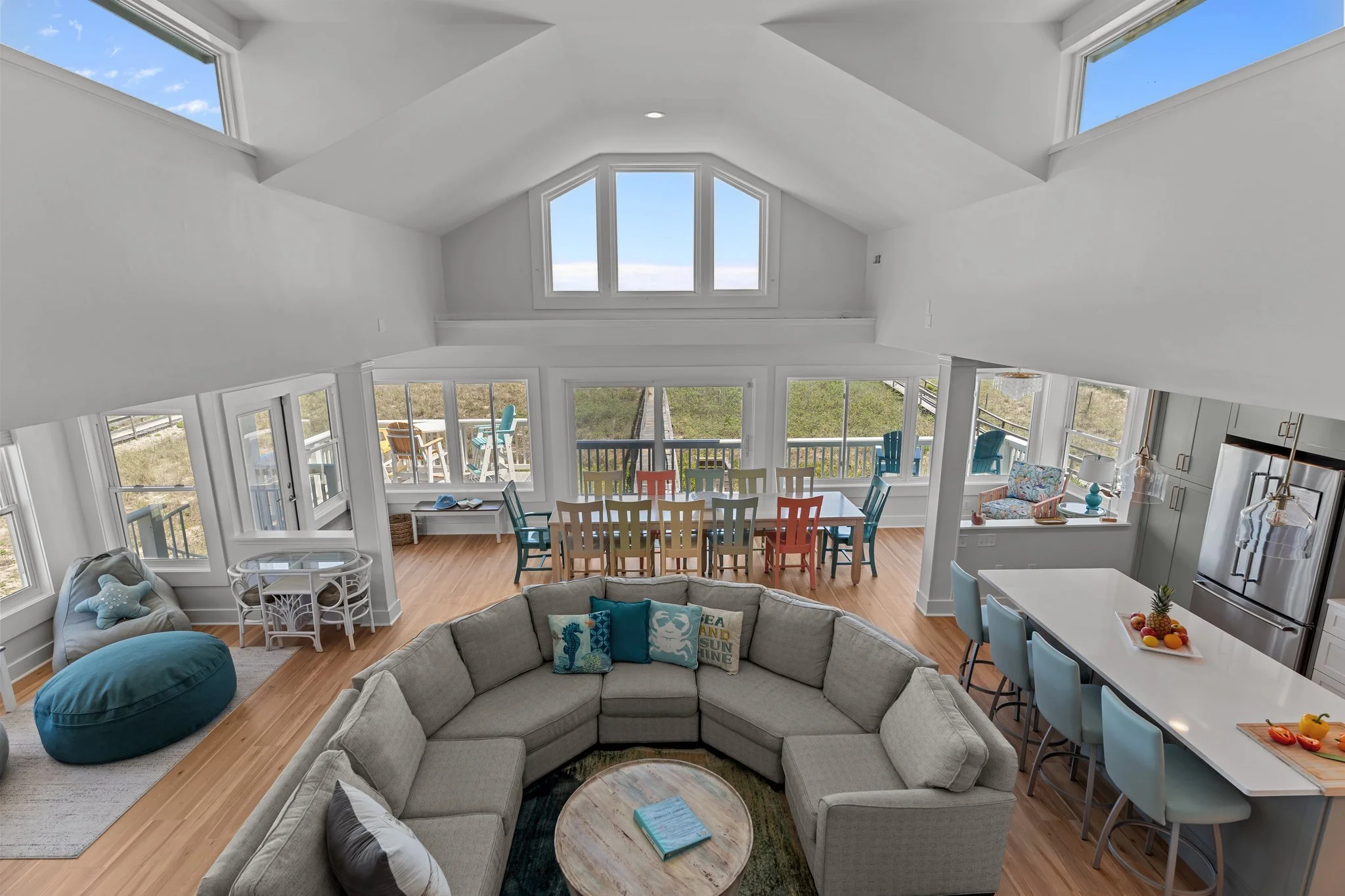A spacious living room with large windows, a vaulted ceiling with skylights, and a dining area with colorful chairs, a sitting area with a sectional sofa, and a view of a beach outdoors.
