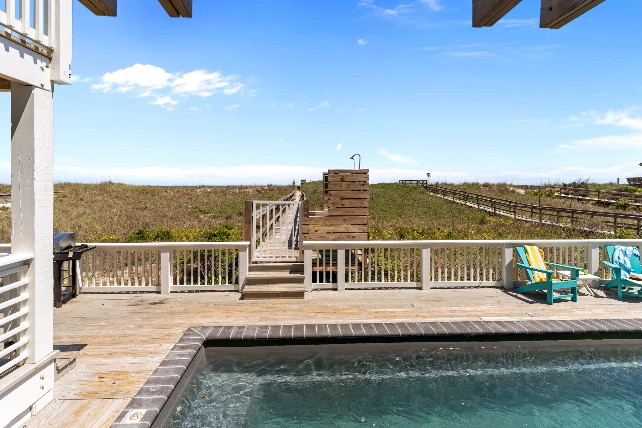 Part view of a backyard pool with a wooden deck, outdoor chairs with striped cushions, and a view of grassy dunes and blue sky with some clouds.
