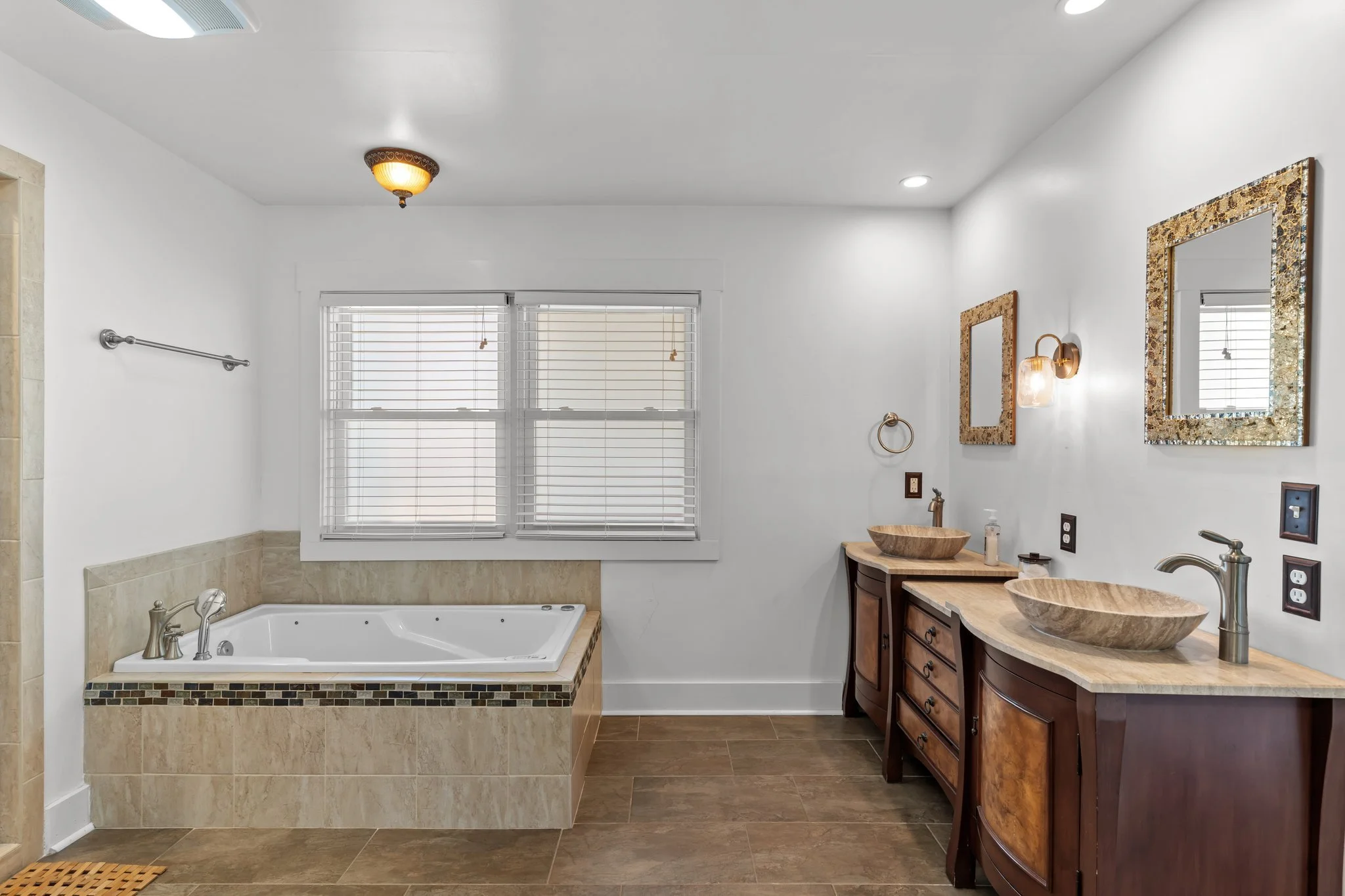 Modern bathroom with a soaking bathtub, dual wooden vanities with vessel sinks, framed mirrors, and a large window with blinds.