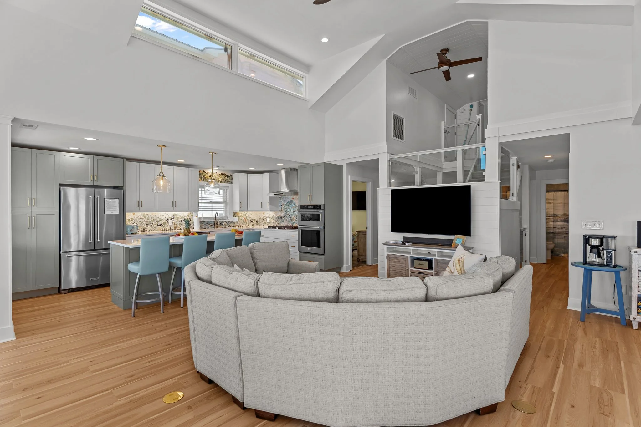 Open-concept living room and kitchen with vaulted ceiling, large window, ceiling fan, and hardwood floors. The living area has a curved sofa, and the kitchen features light gray cabinets, a white island with light blue chairs, stainless steel applian