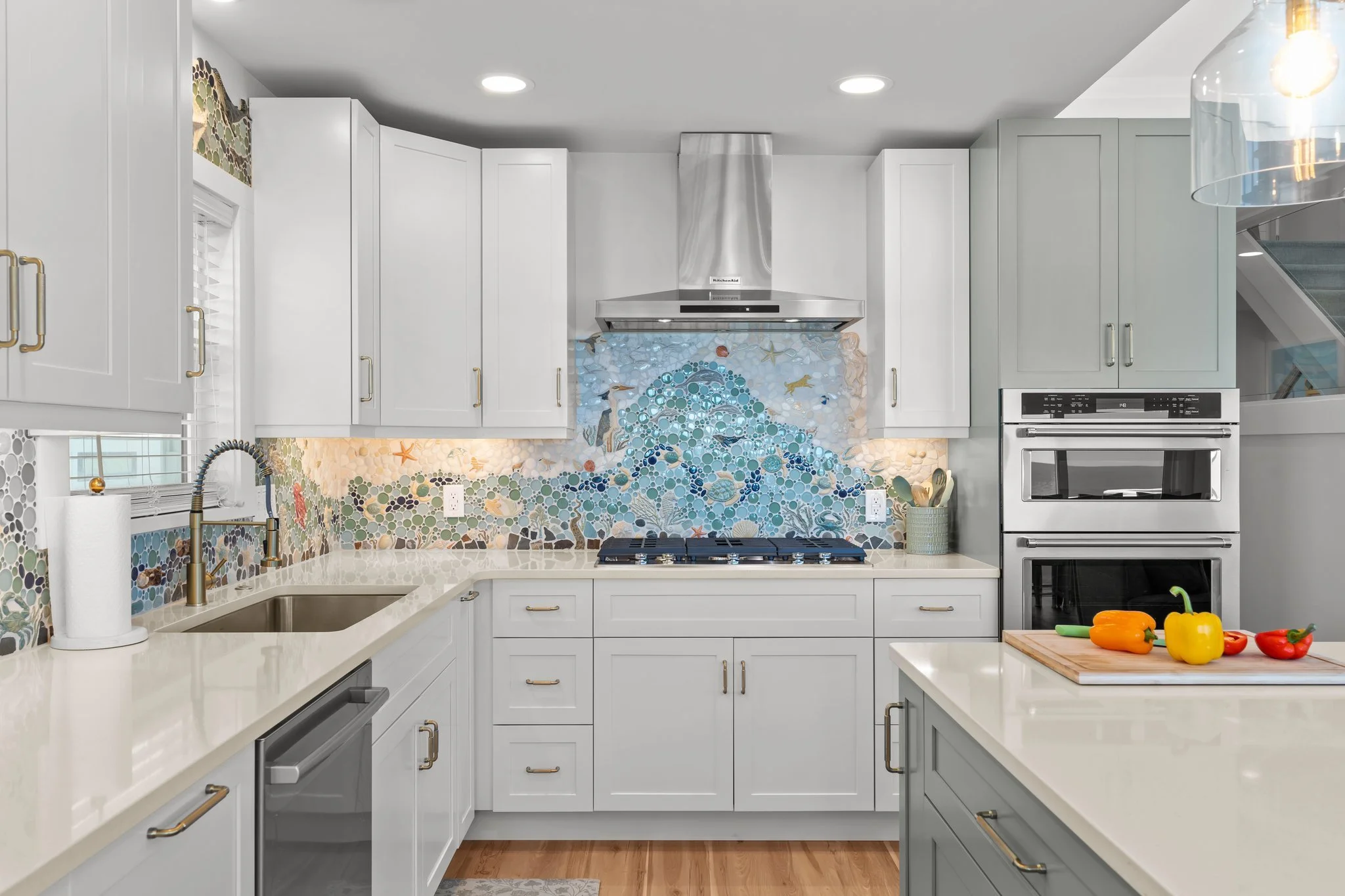 A modern kitchen with white cabinets, a marble countertop, and a mosaic backsplash featuring ocean-themed designs including fish, starfish, and sea turtles. There is a stove and range hood, with colorful peppers on the counter.