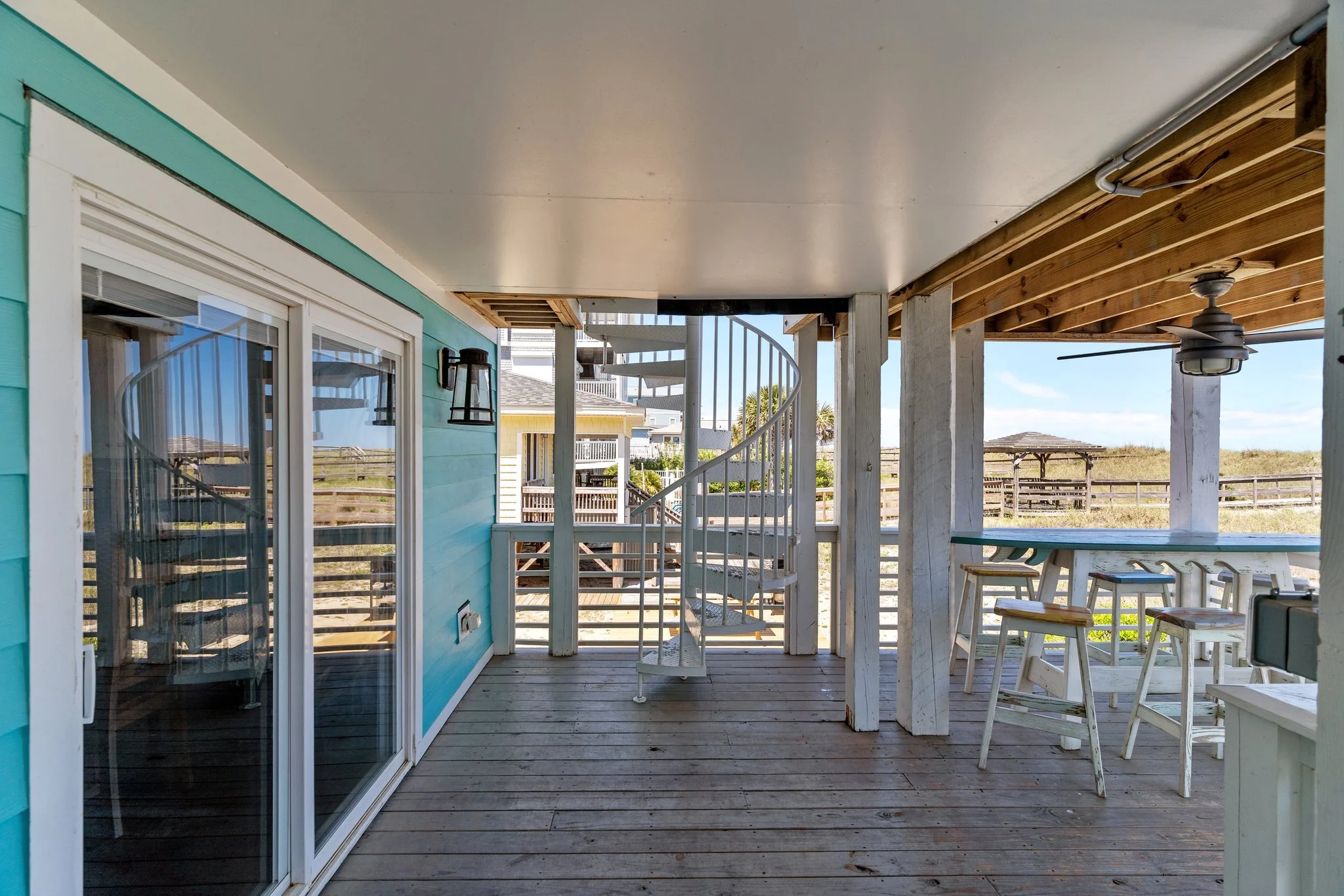 View of a beach house porch with sliding glass doors, wooden flooring, white columns, a spiral staircase leading upward, an outdoor ceiling fan, and a table with chairs, overlooking a sandy beach and distant houses under a partly cloudy sky.