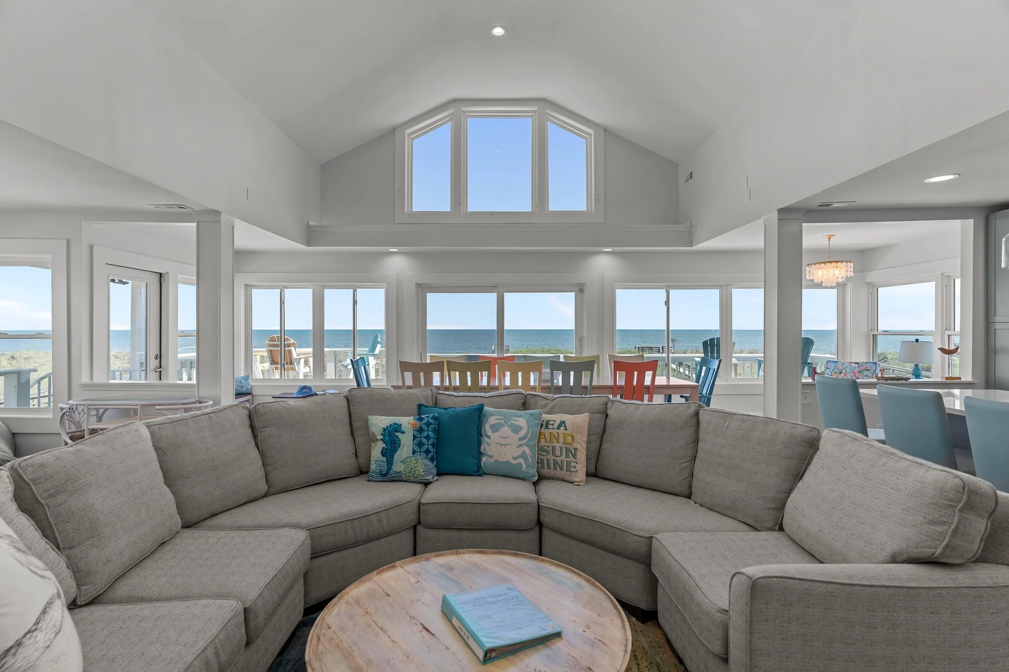 Living room with large windows overlooking the ocean, a beige sectional sofa with decorative pillows, and a round coffee table with a book on it.