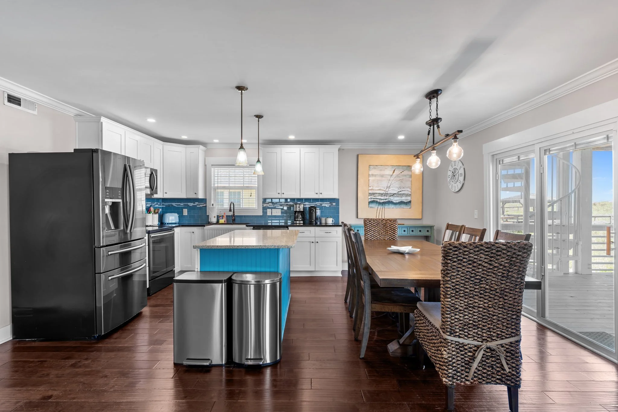 Open-concept kitchen and dining area with white cabinets, stainless steel appliances, a blue kitchen island, hardwood floors, and a wooden dining table with woven chairs. Large windows and a sliding glass door let in natural light.