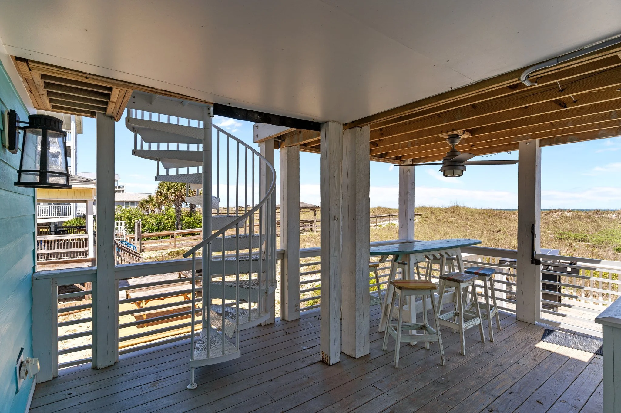 View of a beachside outdoor patio with seating, a bar table with stools, a ceiling fan, and a spiral staircase leading to the upper level, overlooking grassy dunes and a clear sky in the background.