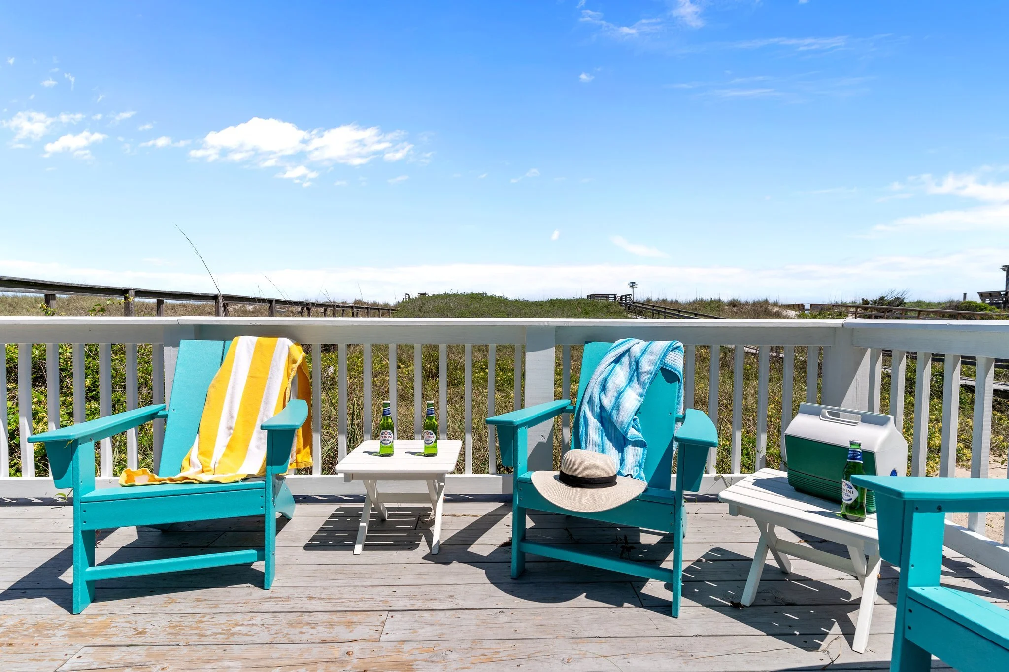 Beachside deck with blue chairs and towels, a sun hat, a small white table, and bottles of soda, overlooking grassy dunes and a blue sky.