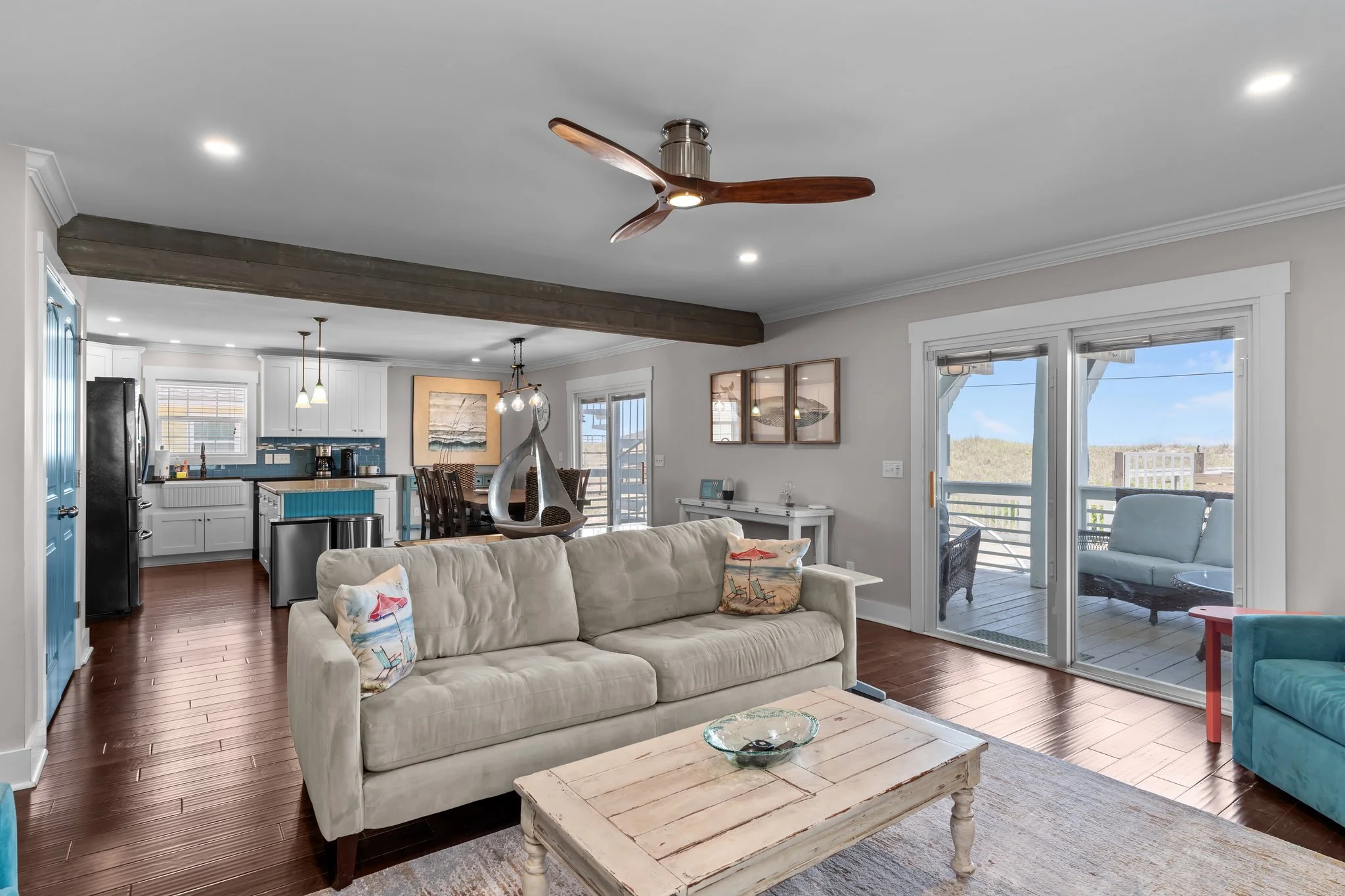 Living room with beige sofa, wooden coffee table, and sliding glass door leading to outdoor deck. Kitchen in background with white cabinets, blue backsplash, and hanging pendant lights. Ceiling fan with wooden blades is visible. Decor includes coasta
