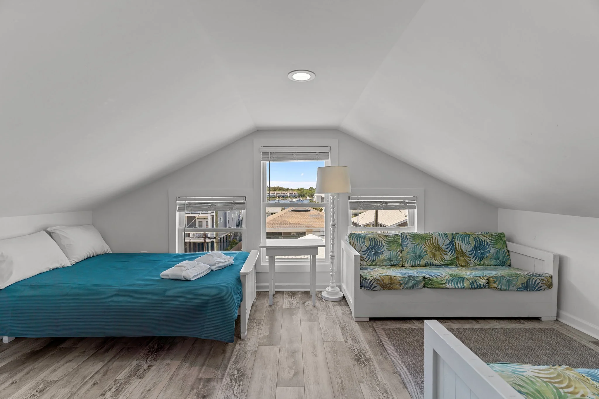 Attic bedroom with white walls, sloped ceiling, large window, bed with blue bedding, tropical print loveseat, and small white table with a lamp near the window.