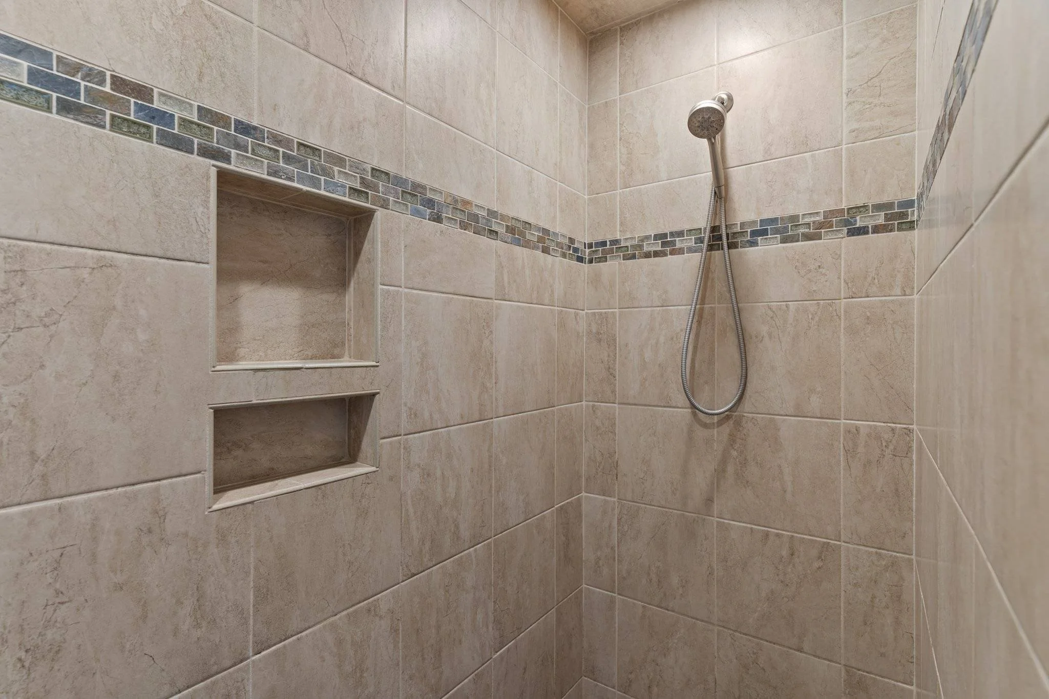 A tiled shower with built-in niche, beige tiles, a decorative tile border, and a handheld showerhead.