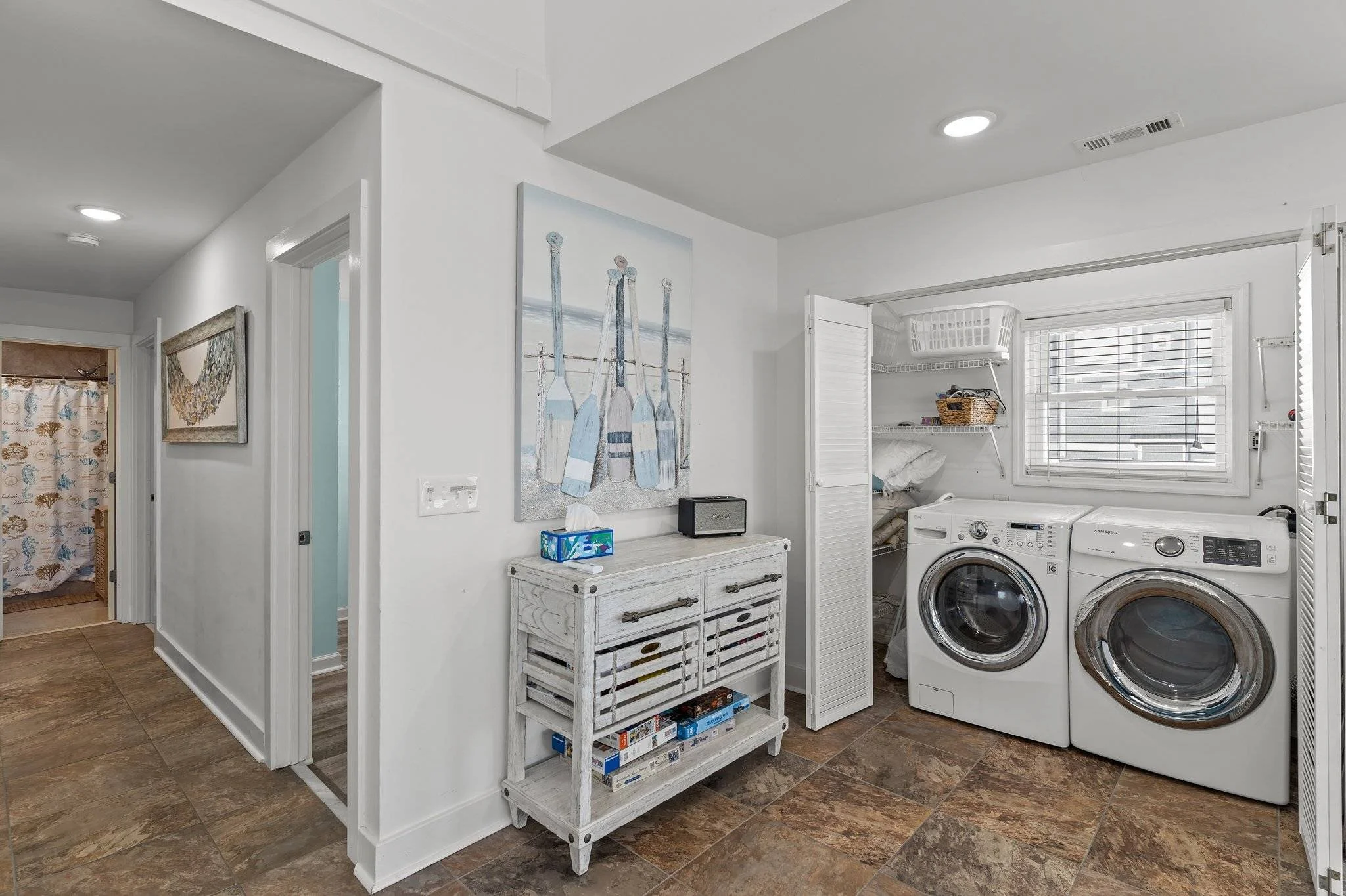 A laundry room with white walls and a window with blinds. There are two front-loading washers, a white washing machine, and a white dryer. A small white distressed table holds a tissue box and a small black speaker. Above the table is a colorful pain