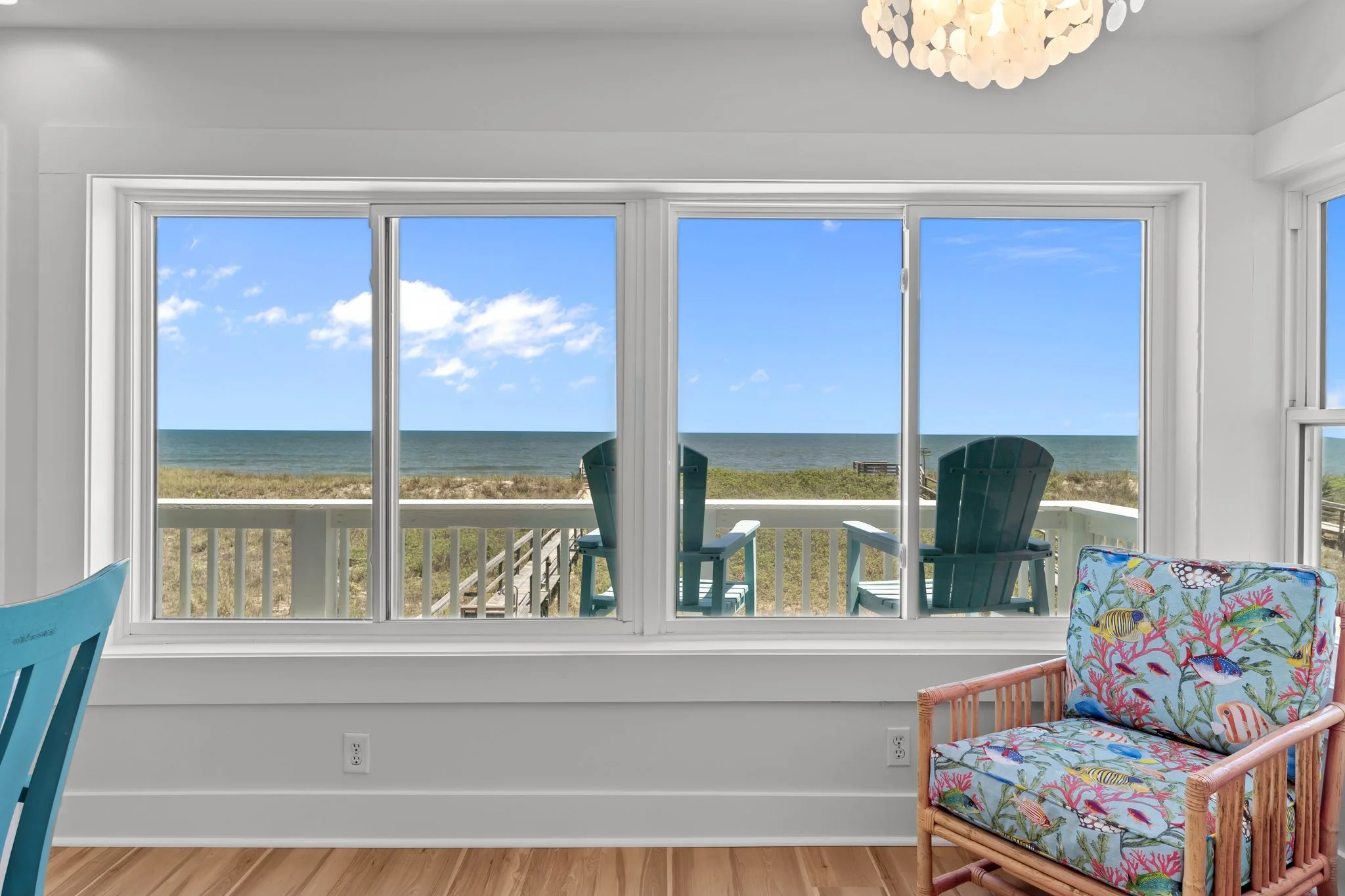 View through large sliding glass door to a sandy beach with ocean waves and a clear blue sky. Inside, there are blue chairs on the porch, and part of a colorful cushioned chair with a coral and fish pattern is visible in the foreground.