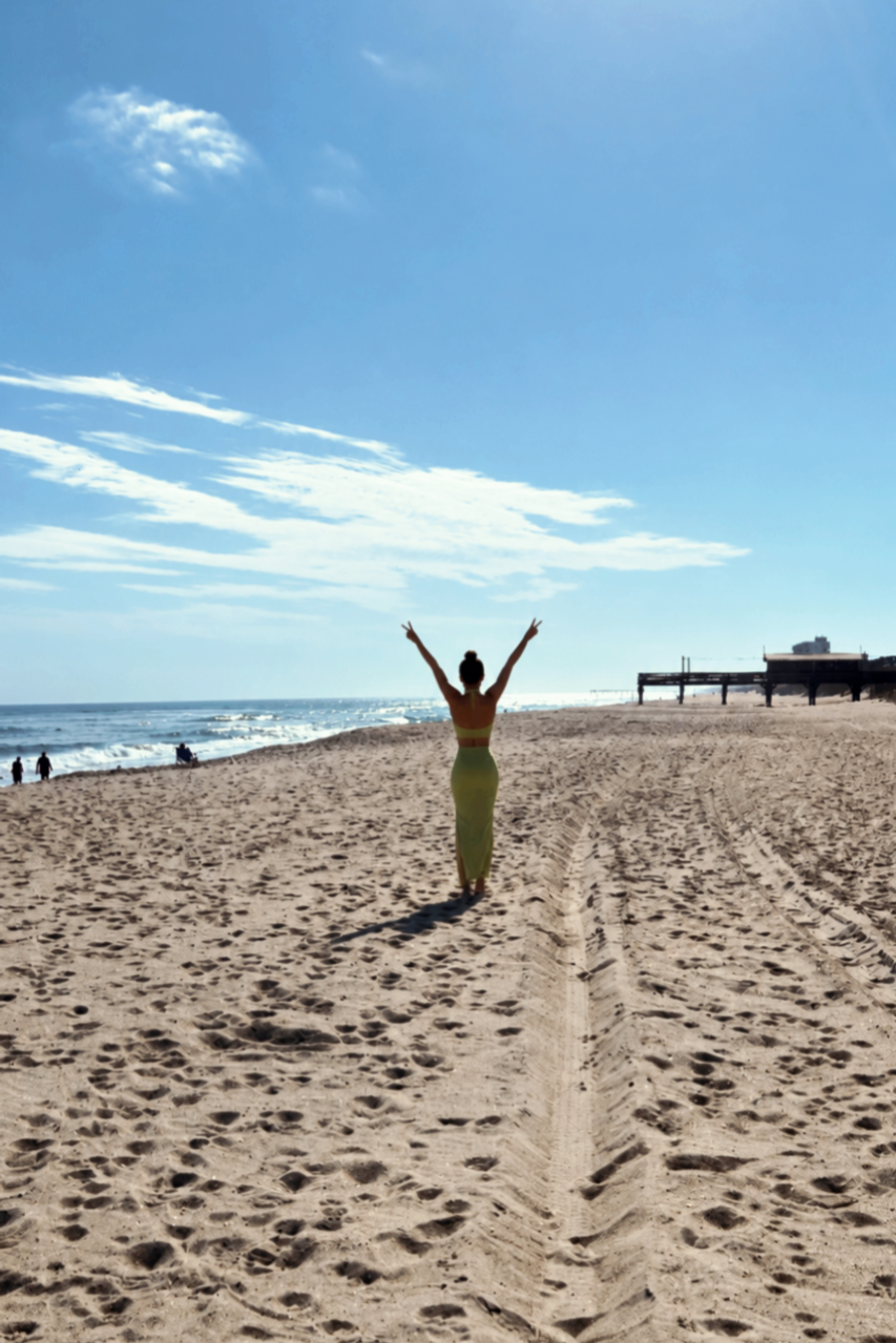 Woman walking on a sandy beach with arms raised, ocean waves in the background, clear blue sky, some clouds, and distant people and buildings.