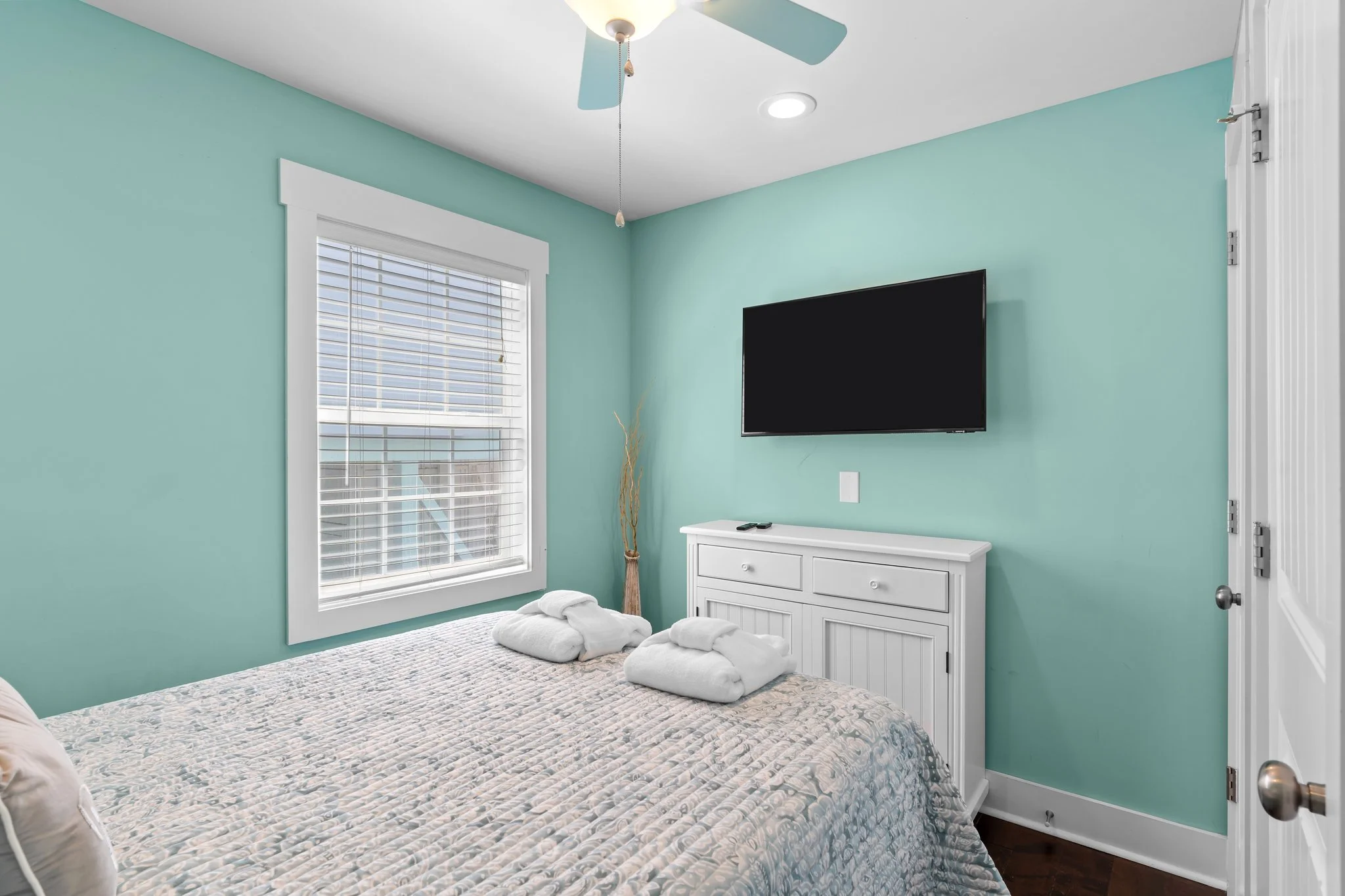 A bedroom with green walls, a window with white blinds, a wall-mounted flat-screen TV, a white dresser, and a bed with a patterned quilt and pillows. Towels are folded on the bed, and a ceiling fan is visible.