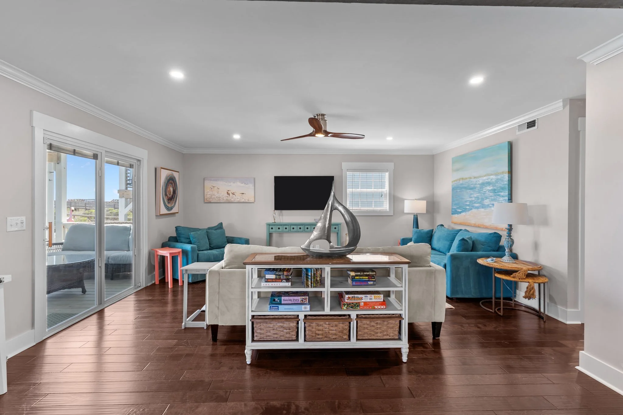 A living room with a white sofa, blue armchairs, and a coffee table filled with books. There is a fireplace with a decorative sculpture on the table. The room has wooden flooring, artwork on the walls, and a ceiling fan. There's a sliding glass door 