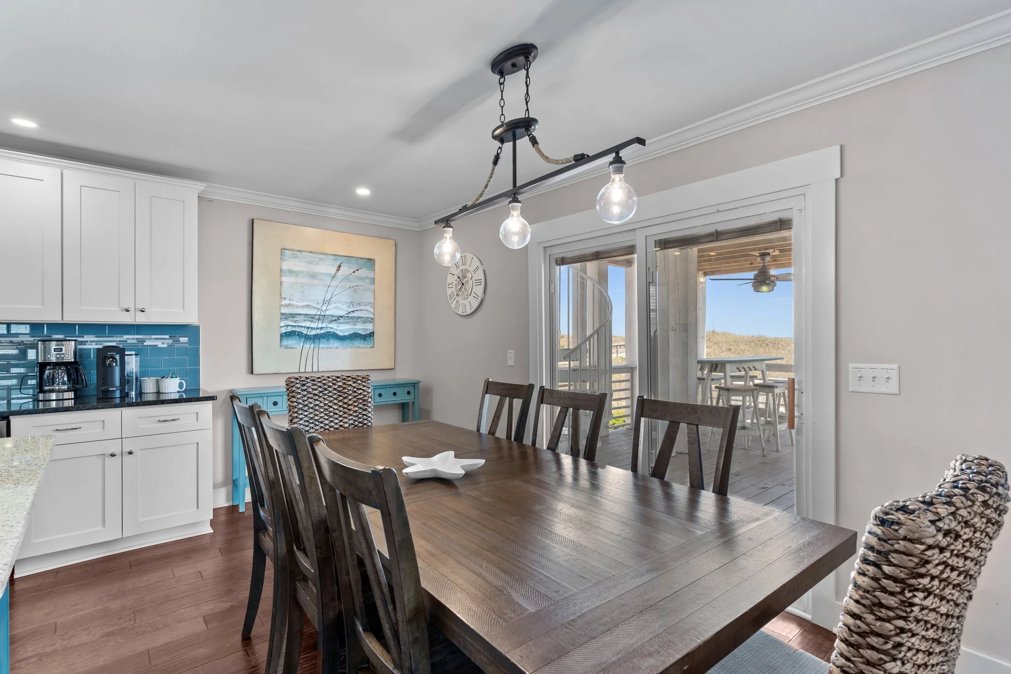 Interior view of a dining area with a wooden table surrounded by chairs, a hanging light fixture with three exposed light bulbs, white kitchen cabinets, a blue sideboard, a wall clock, and a sliding glass door leading to an outdoor patio with a ceili