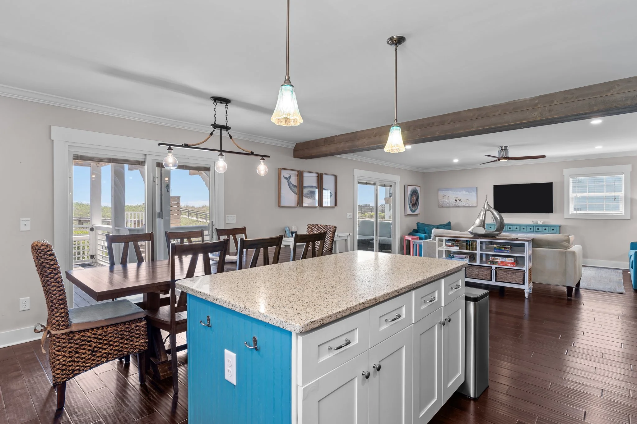 Open-concept living and dining room with a kitchen island, wooden dining table with chairs, windows, artwork on the walls, a TV, and a ceiling fan.