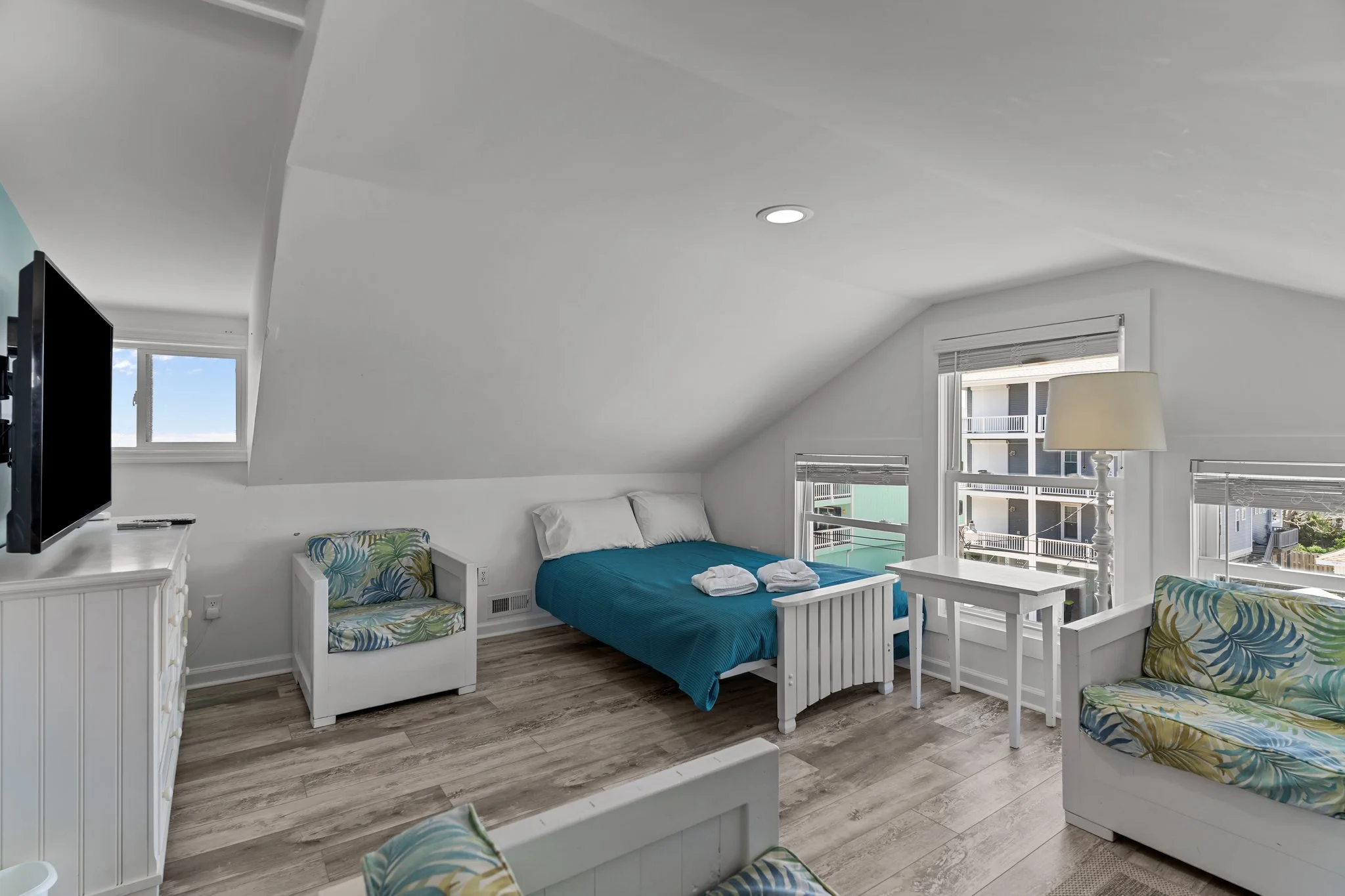 Bright bedroom with white walls, sloped ceiling, and multiple windows showing buildings outside. Features a blue bed, white furniture, a TV, and colorful armchairs.