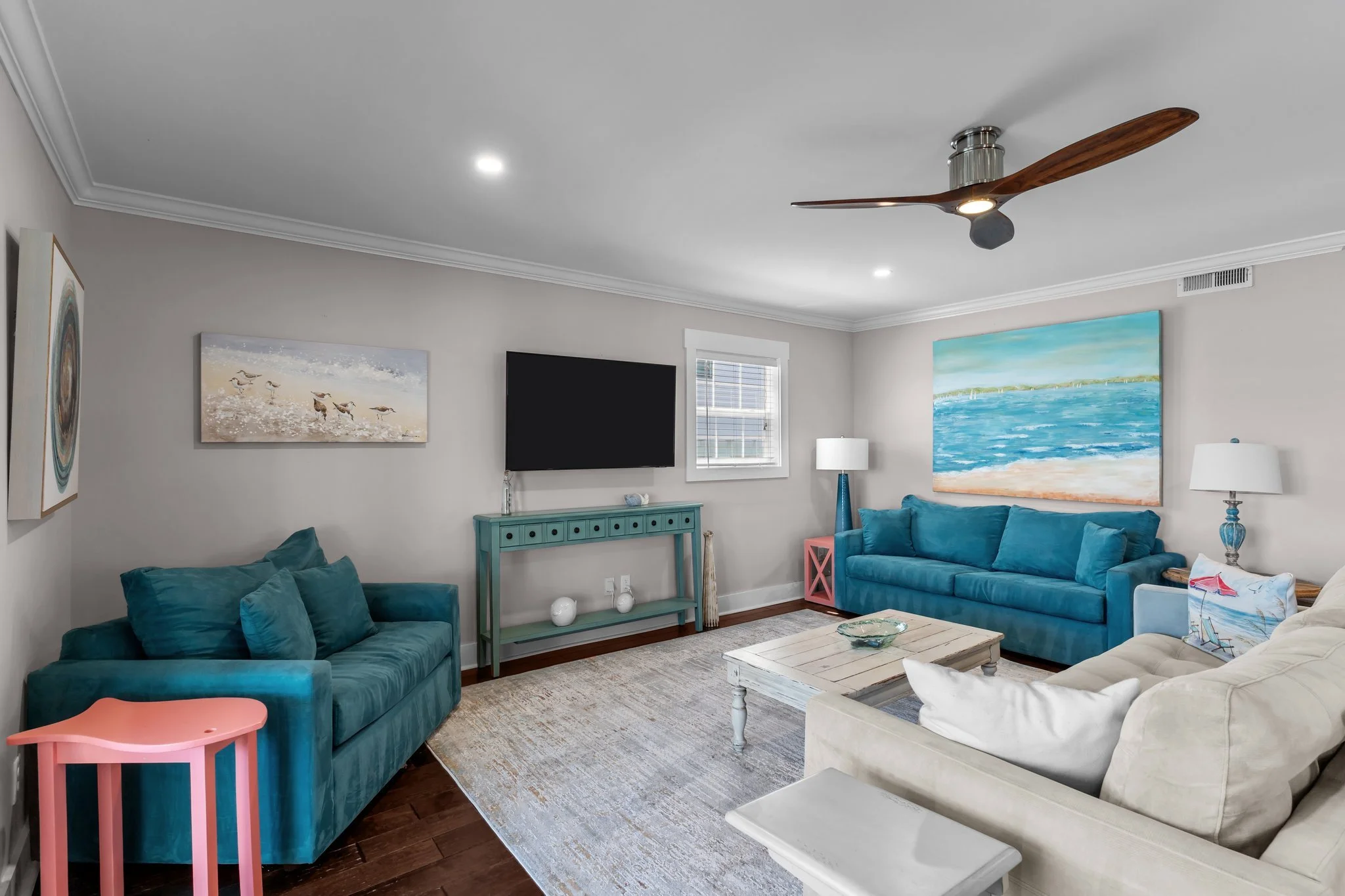 Living room with gray painted walls, turquoise velvet sofas, a white coffee table, a beige armchair, wall paintings of seagulls and ocean, a flat screen TV, a window with blinds, a turquoise side table with decorative objects, a floor lamp, and a cei