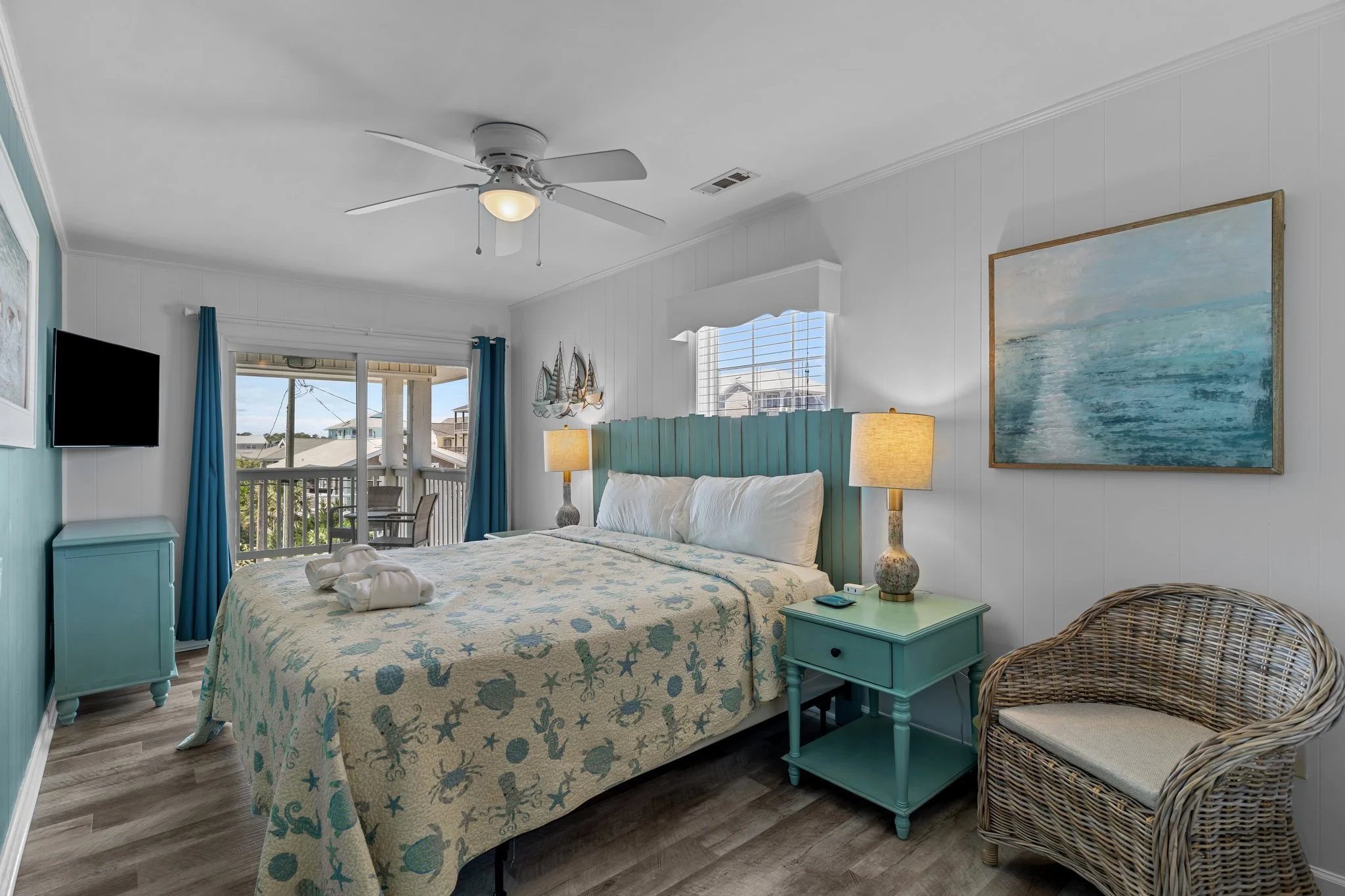 A bedroom with a white ceiling fan, teal headboard, large window with blue curtains, and a wall-mounted flat-screen TV. There is a bed with a seashell and starfish patterned bedspread, bedside table with a lamp, and wicker chair.