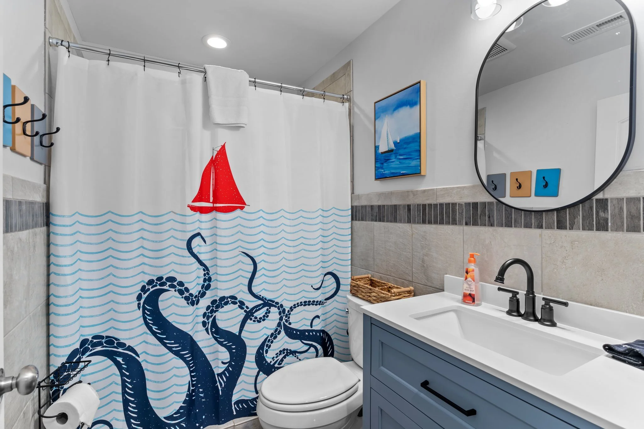 A bathroom with a nautical theme. It features a shower curtain with a red sailboat and blue waves, a framed picture of sailboats on the wall, and a sink with a blue cabinet, black faucet, and soap dispenser. There are hooks on the wall, a basket belo