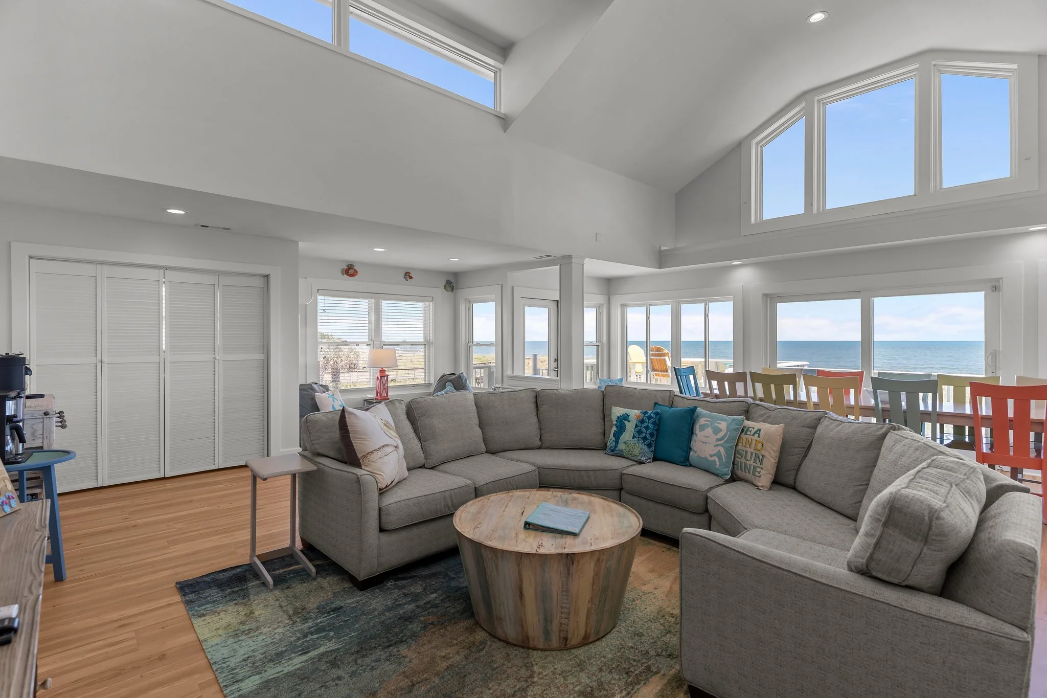 Living room with large sectional sofa, window view of the ocean, and colorful chairs in the dining area.