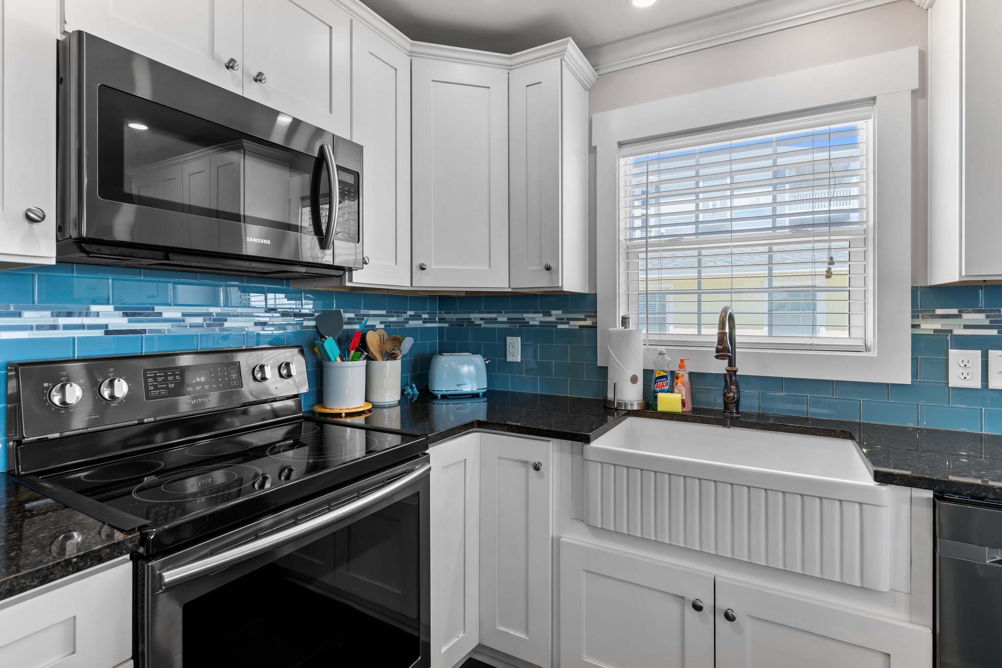A modern kitchen with white cabinets, a stainless steel microwave, a black stove, a blue tile backsplash, and a window with white blinds.