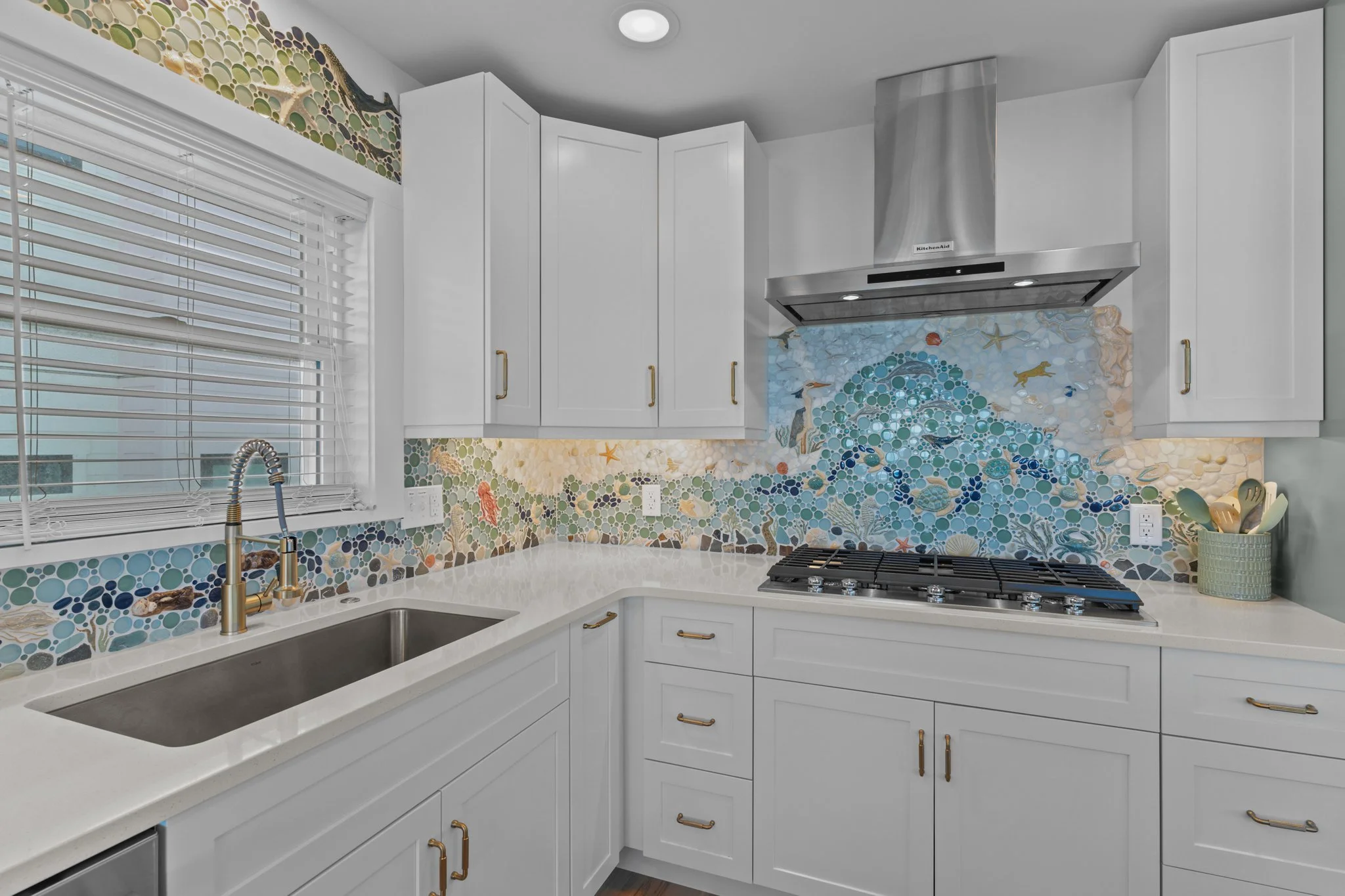 A modern white kitchen with a mosaic tile backsplash depicting an ocean scene with fish, starfish, and seaweed, stainless steel range hood, and gold handles on cabinets. A window with blinds is above the sink, and there is a utensil holder on the cou