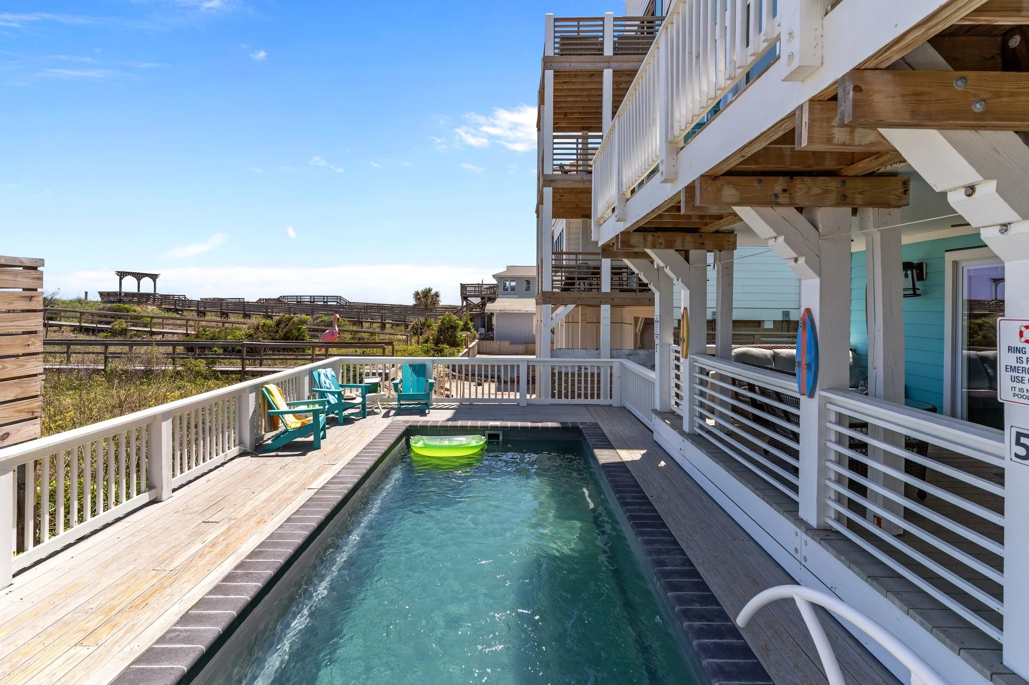 A backyard with a small pool, turquoise Adirondack chairs, and a multi-level wooden deck with a white railing, overlooking beach dunes with a pink flamingo statue and a gazebo in the distance.