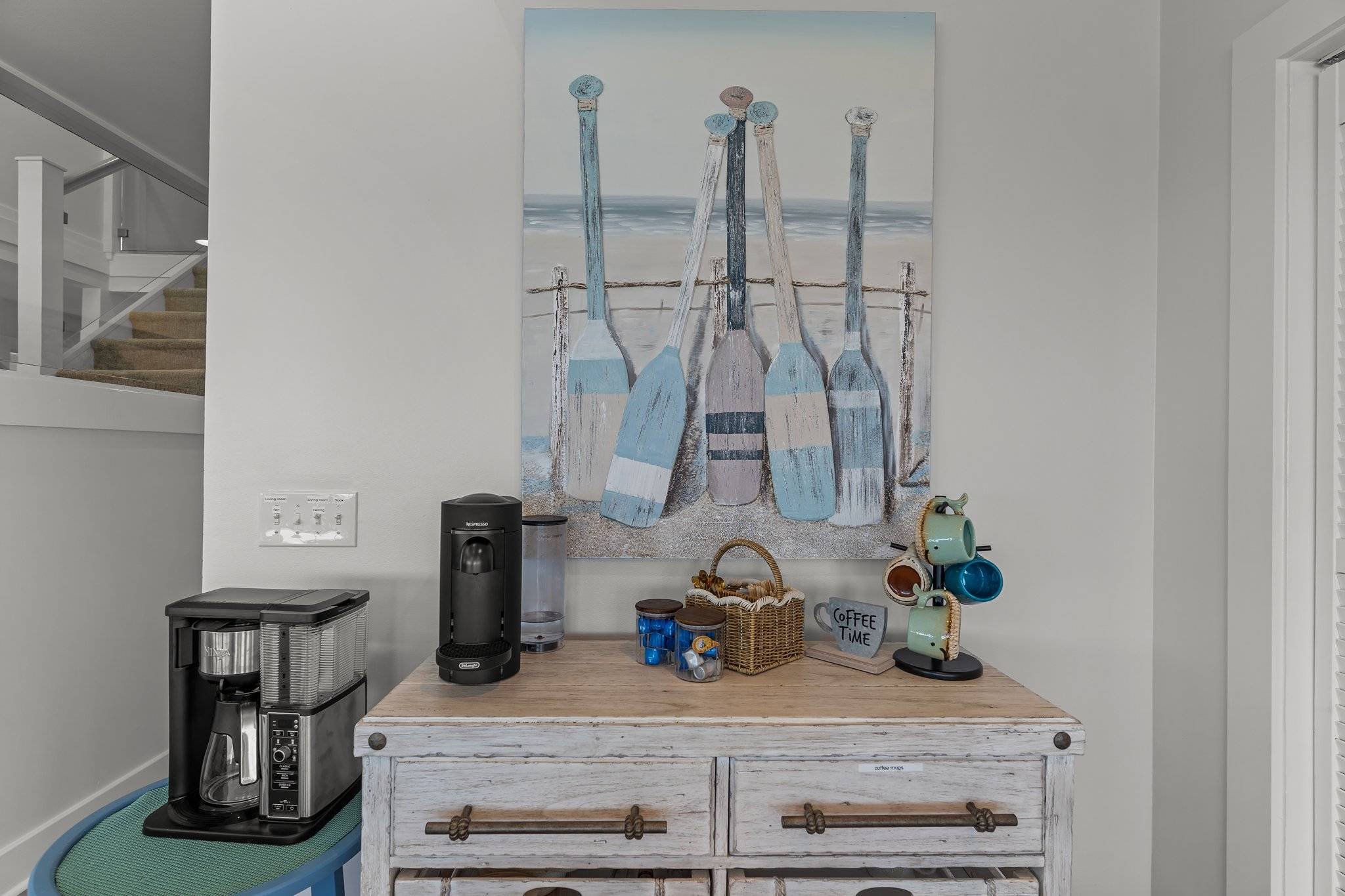 A kitchen countertop with coffee-making appliances, baskets, and mugs, against a white wall with a large beach-themed painting of bottles.