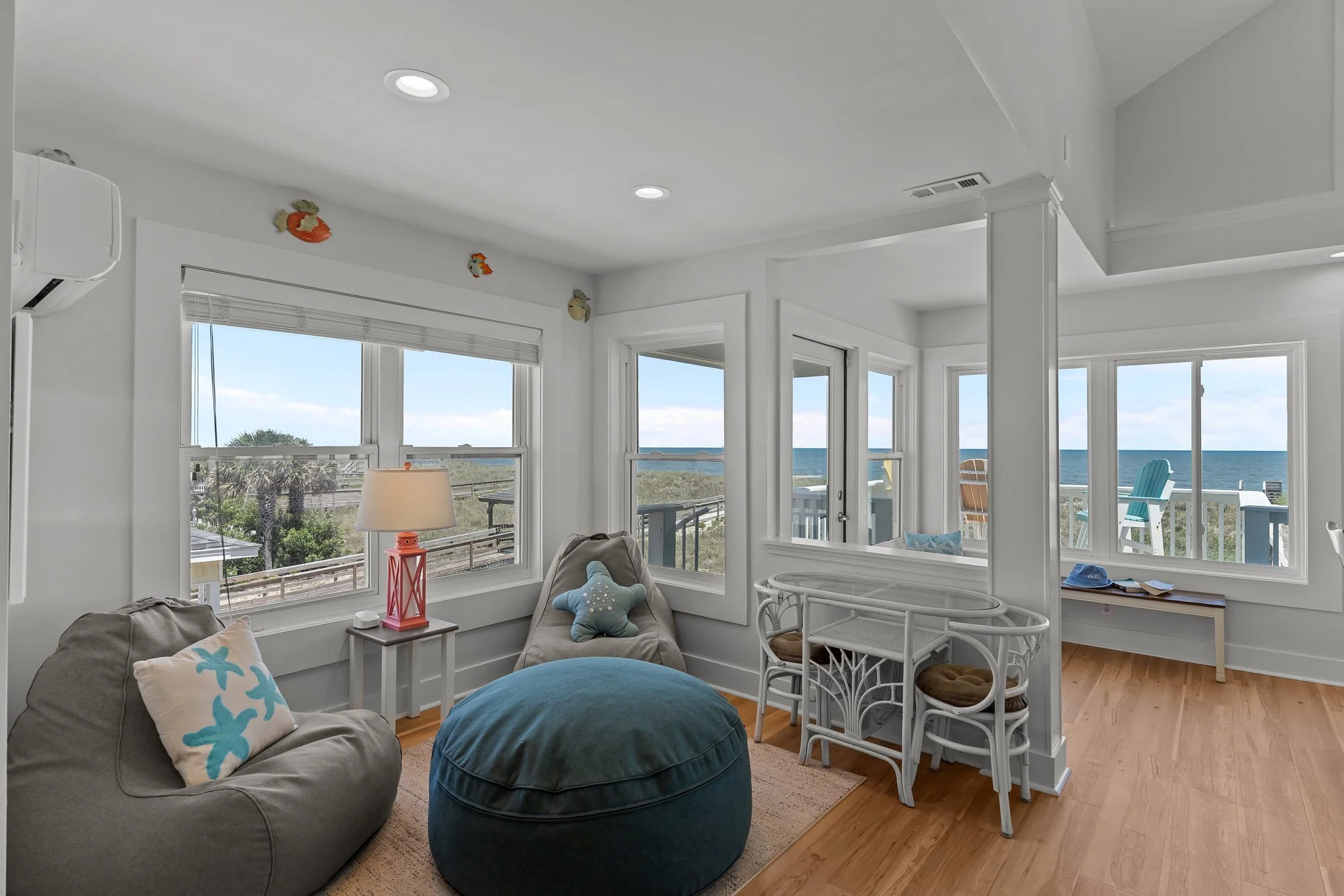 Living room with large windows showing a beach view, furnished with a gray sofa, blue pillows, and a starfish pillow, also includes a round blue ottoman, white wicker chairs, a side table with a pink lamp, and decorative fish wall hangings.